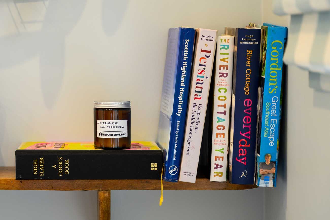 A small wooden shelf featuring a selection of colourful cookbooks, topped with a small amber glass jar candle.