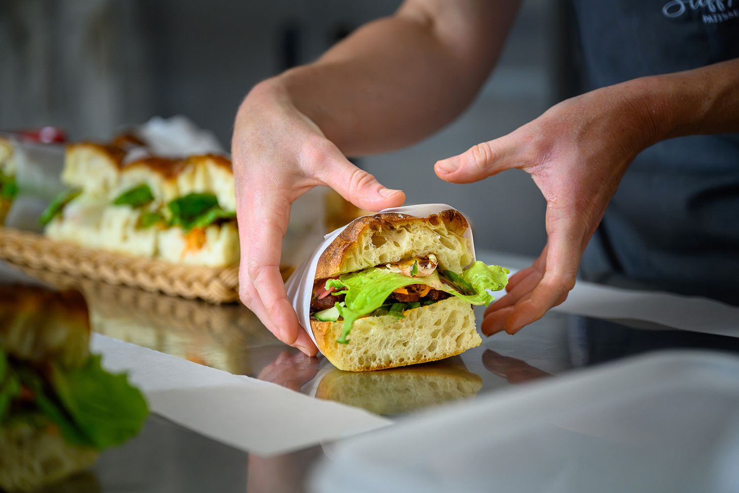 A person's hands holding a freshly prepared sandwich on thick, crusty bread wrapped in white parchment paper on a stainless steel counter.