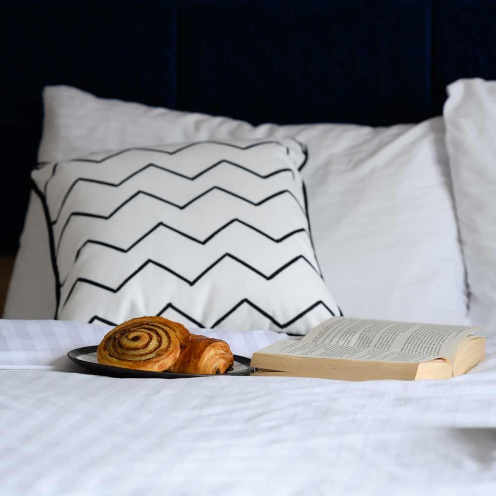 A cinnamon swirl pastry on a plate next to an open book on a bed with white linens and a chevron patterned accent pillow.