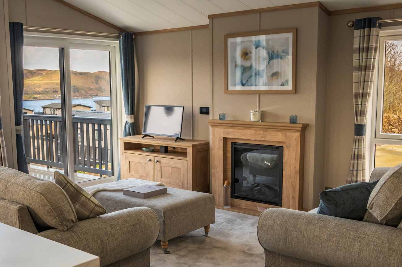 A cosy holiday home lounge with fabric armchairs, an electric fireplace, a TV on a wooden stand, and glass doors to a deck.