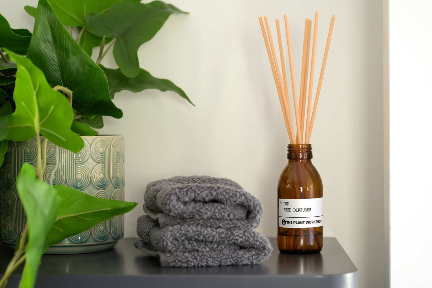 Bathroom shelf with a green houseplant, a stack of folded grey hand towels, and a reed diffuser in an amber glass bottle.
