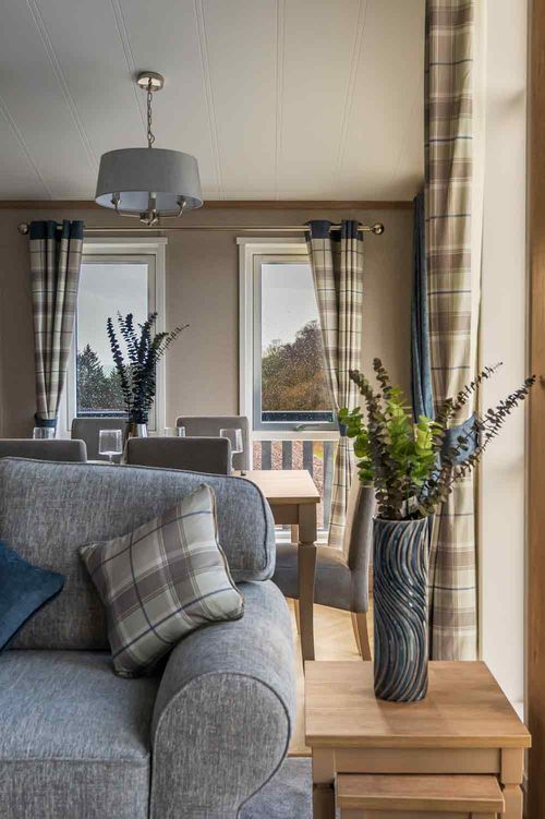 A modern living space featuring a grey sofa with a plaid cushion, a side table with a tall vase, and large windows.