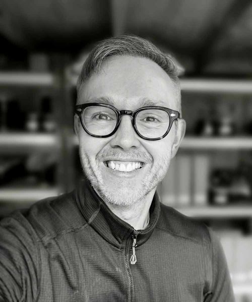 A black and white portrait of Dave owner of Highland Croft Media, wearing a zip-up jacket, with blurred shelving in the background.
