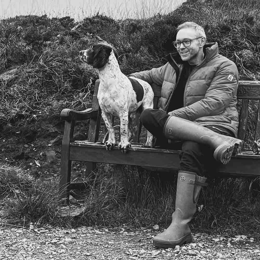 A black and white photo of a smiling man in a winter jacket and wellies sitting on a wooden bench with a spaniel dog.