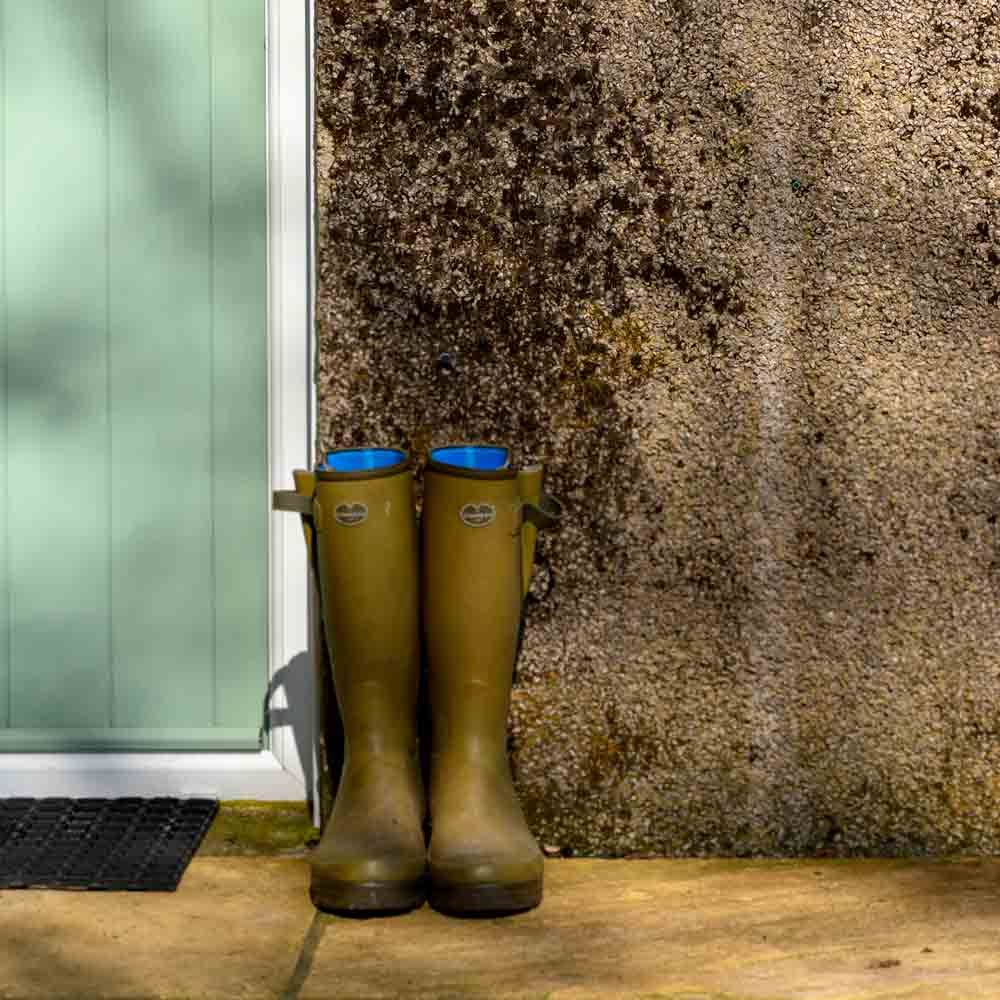 A pair of green rubber wellington boots standing neatly against a textured stone wall next to a light green door frame.