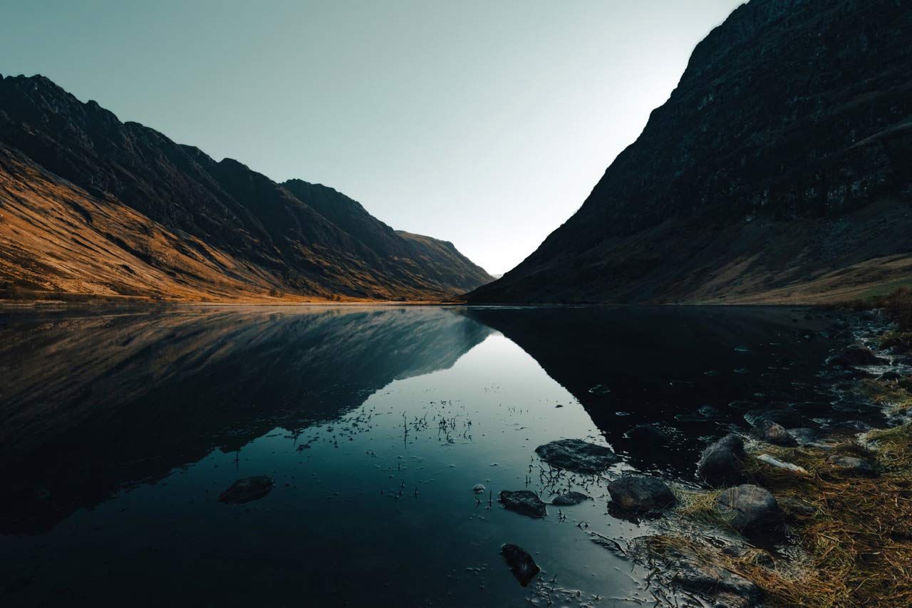 Dramatic Glencoe mountain peaks perfectly reflecting in the calm, dark waters of a loch under a clear sky.