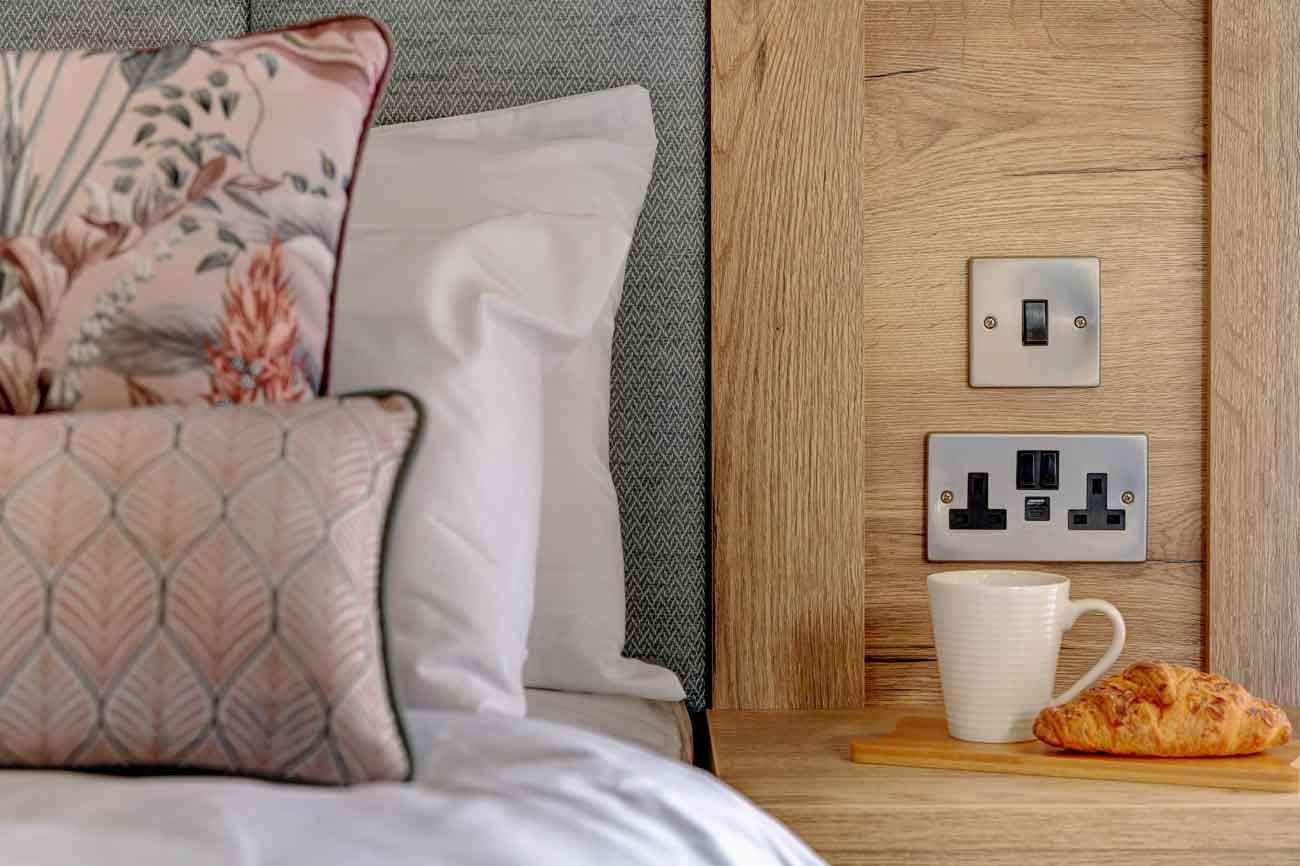 A wooden headboard detail with a white mug and croissant on a built-in shelf next to structured pink patterned pillows.