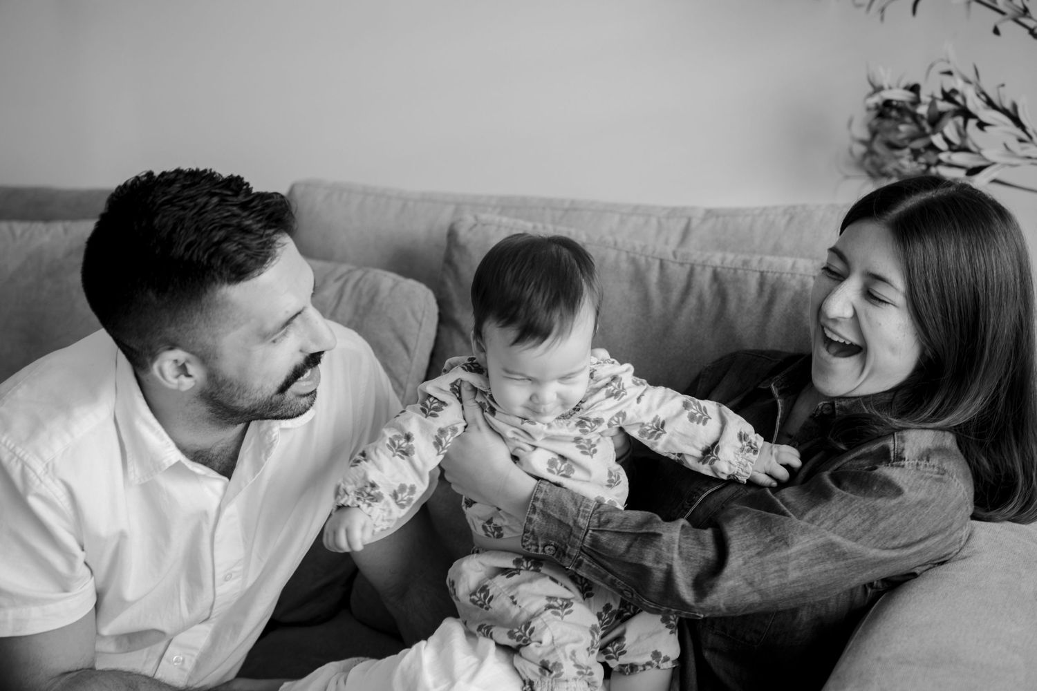 Black and white photo of happy parents sitting on couch laughing with their newborn baby at home.