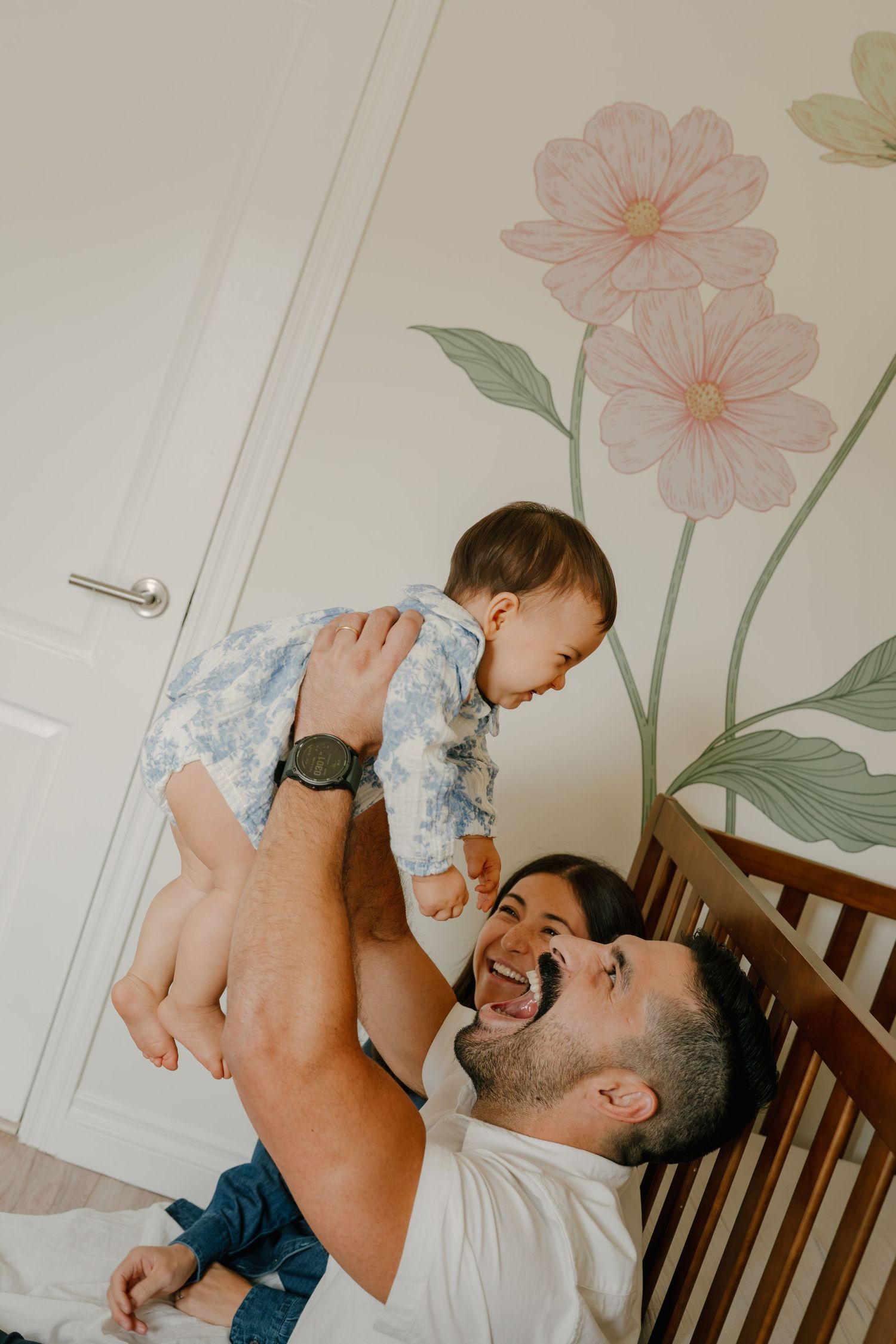 A laughing father lifts his smiling baby in a nursery with a floral wall mural and wooden crib.
