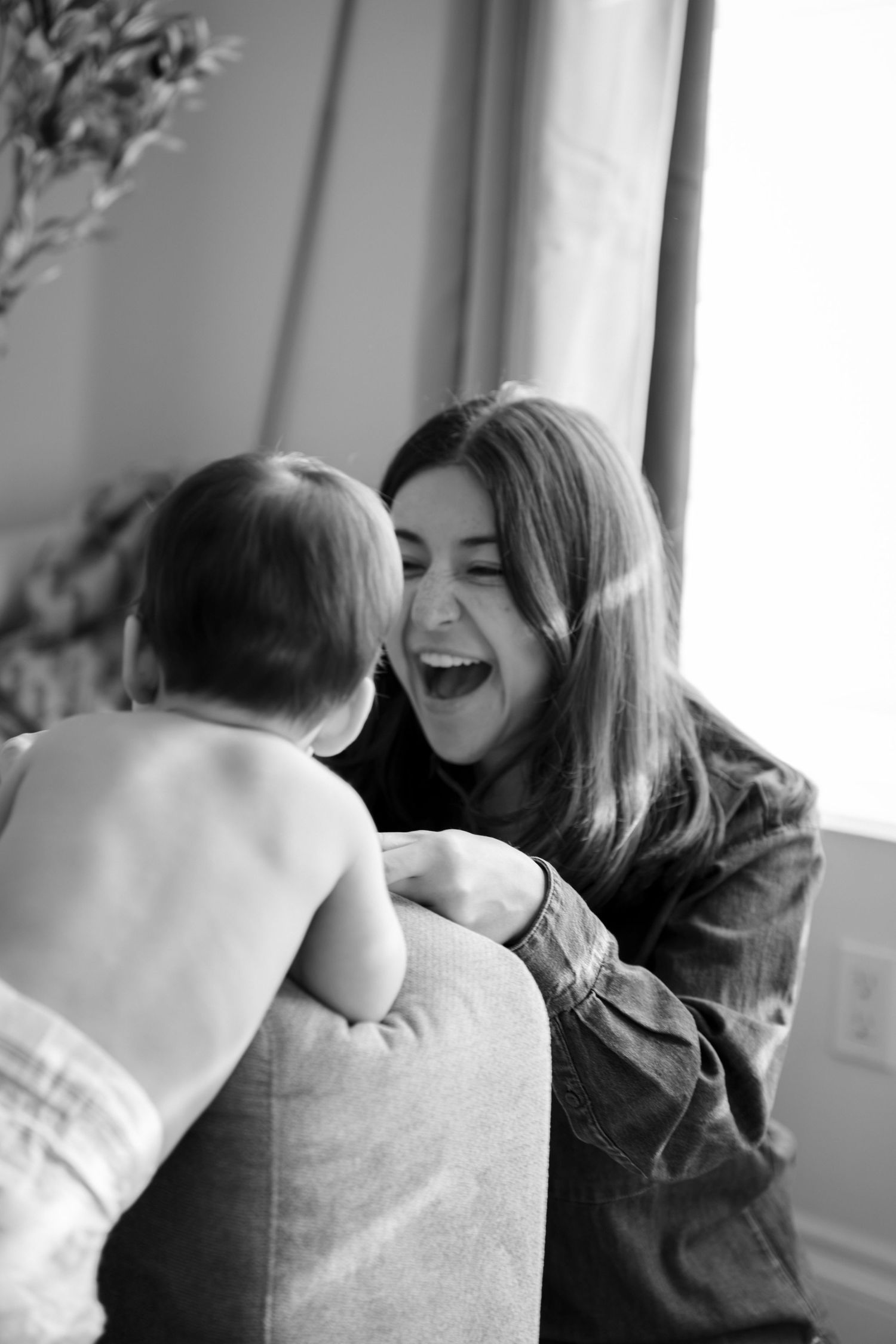 Black and white photo of a laughing young woman leaning in close to a baby sitting on a couch indoors.