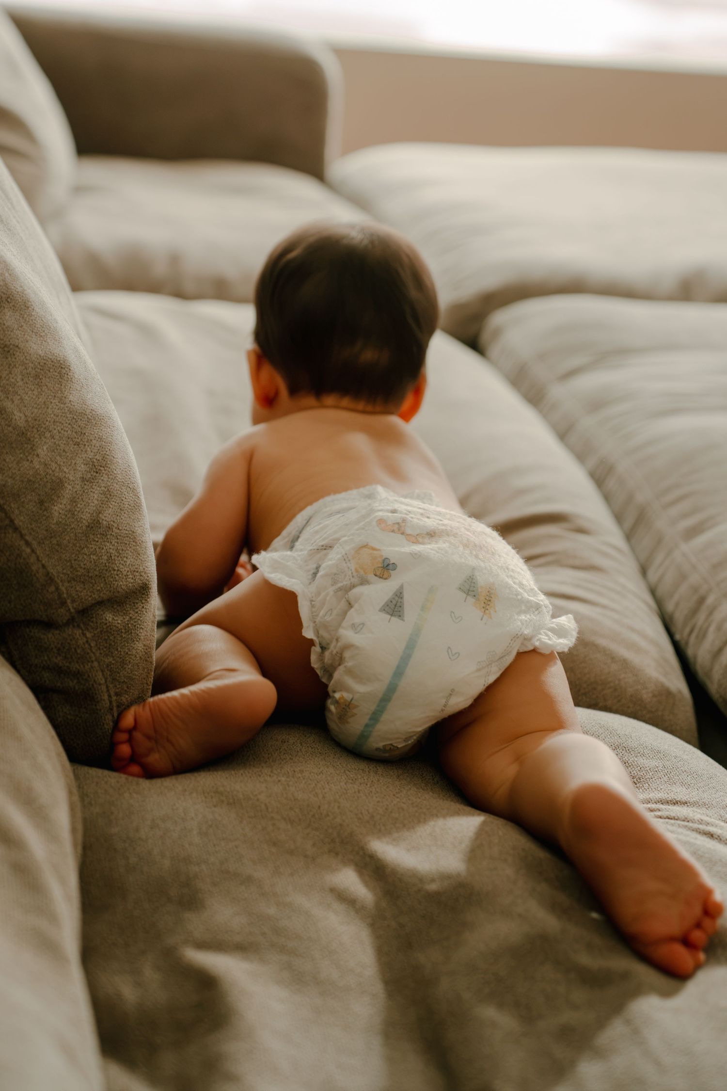 A baby in a white diaper lying on a gray couch, viewed from behind.