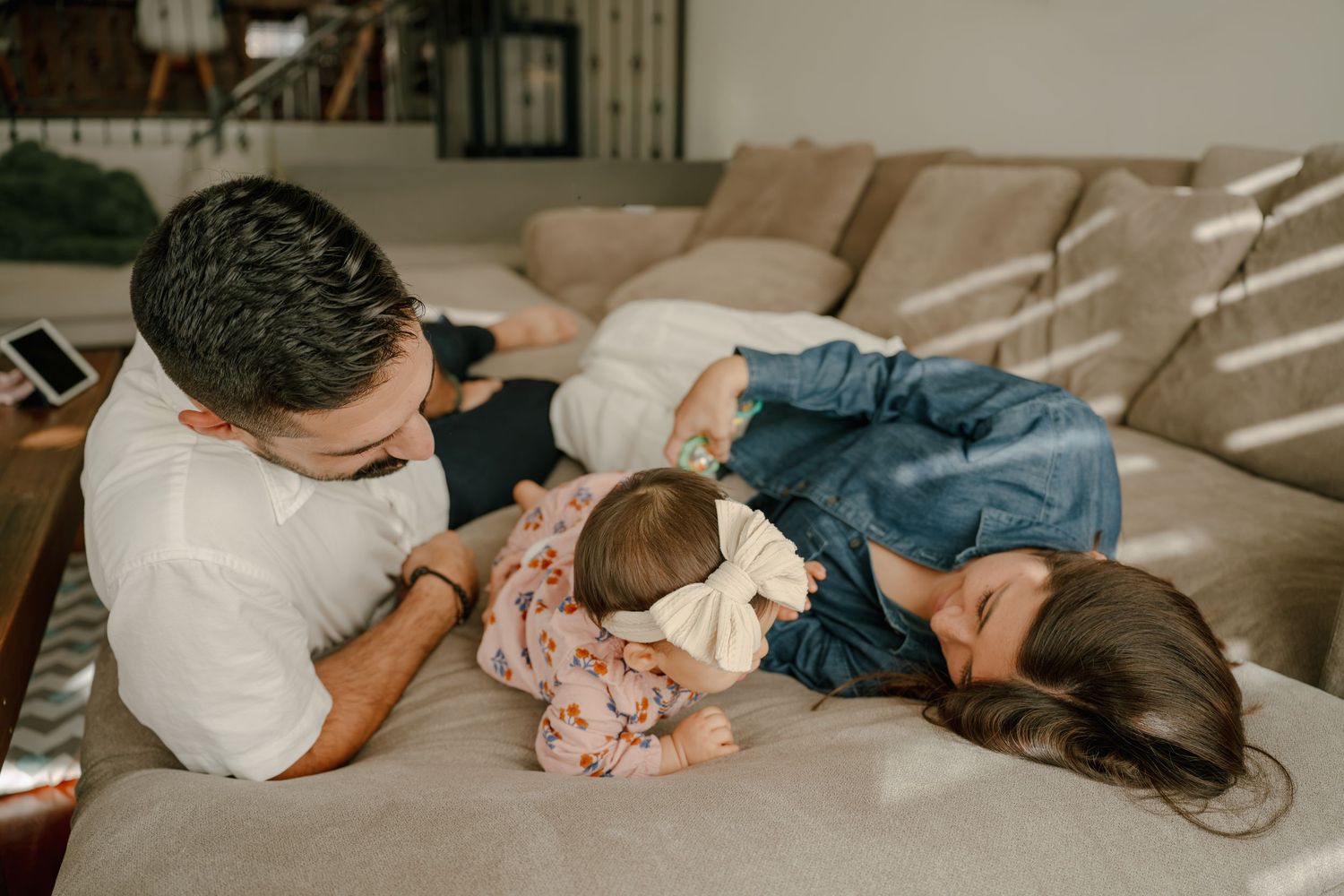 Family with newborn baby bonding on couch, parents lovingly gazing at infant with white bow headband.