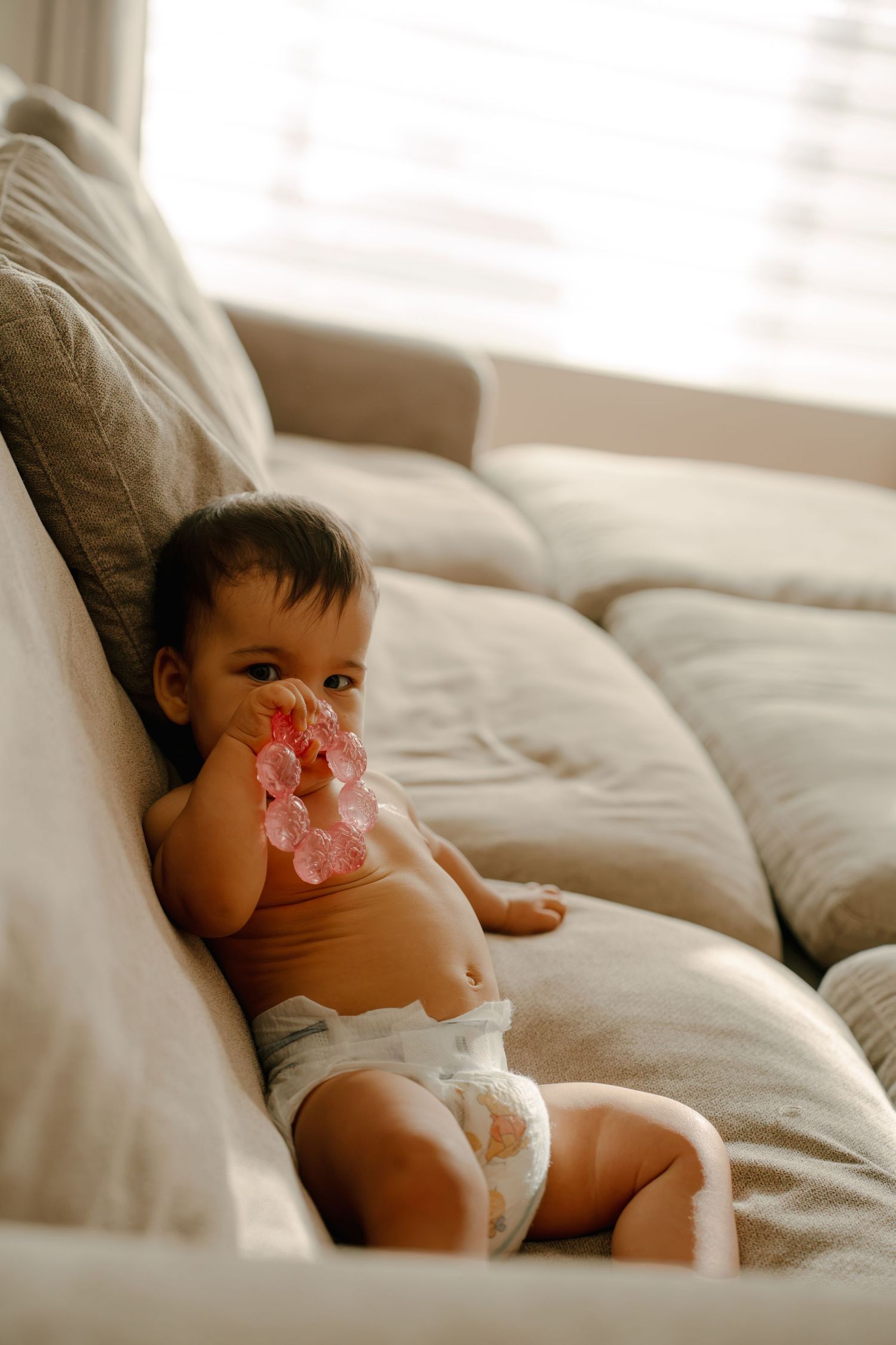 Cute baby in diaper sitting on gray couch chewing pink teething toy in bright living room.
