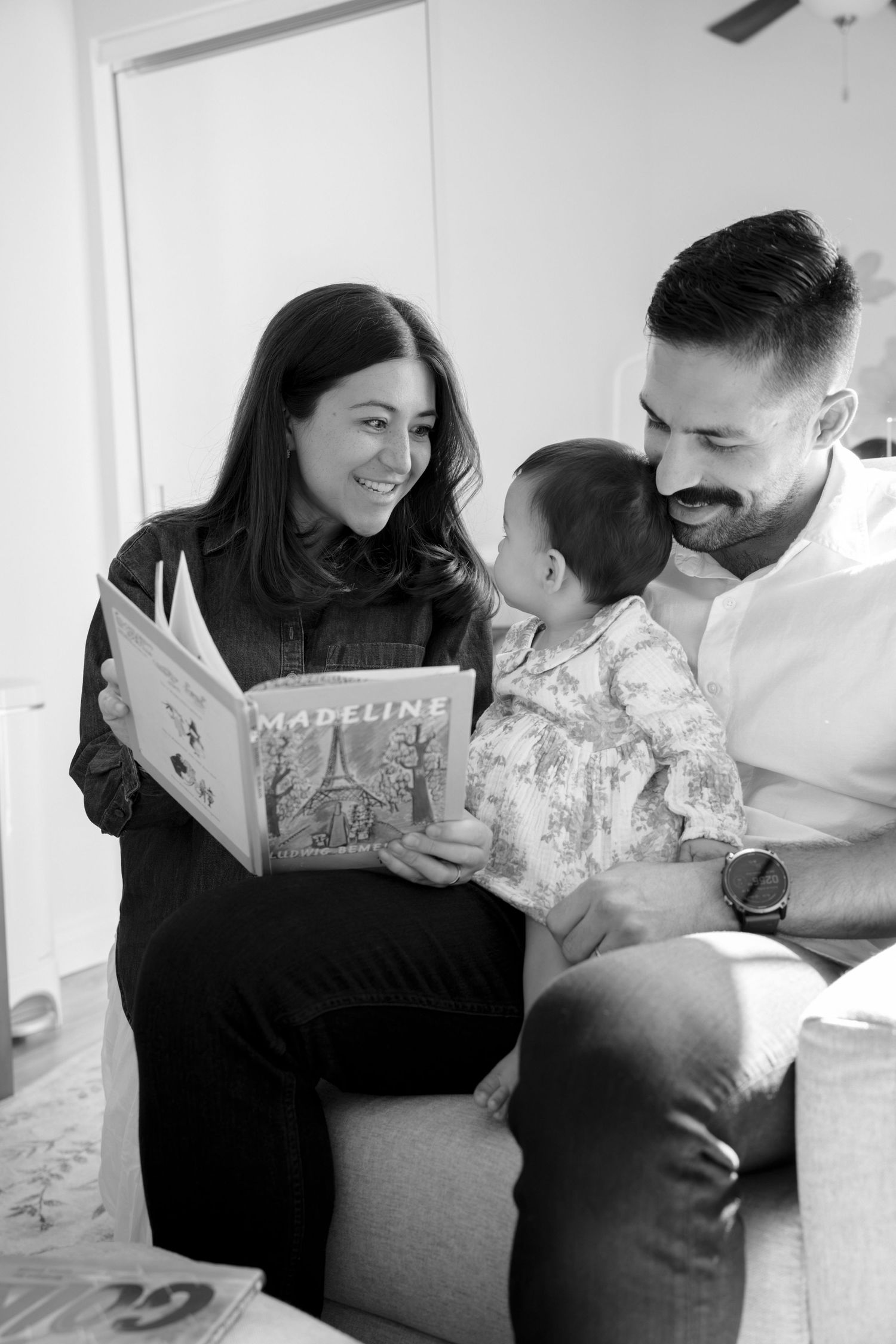 Black and white photo of a couple smiling at their baby while reading the children's book Madeline together.