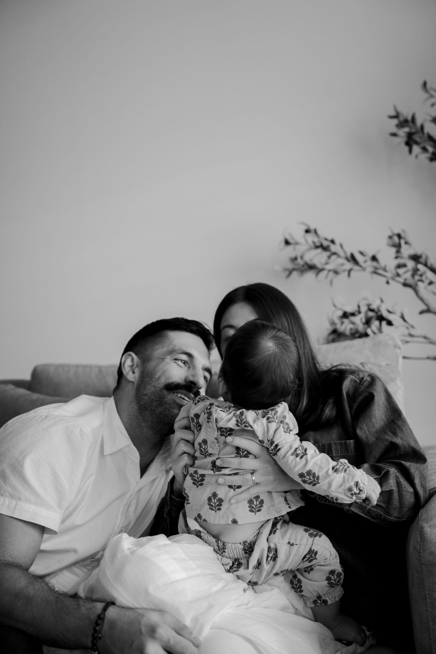 Black and white photo of smiling parents sitting on a couch, lovingly holding their baby in patterned pajamas.