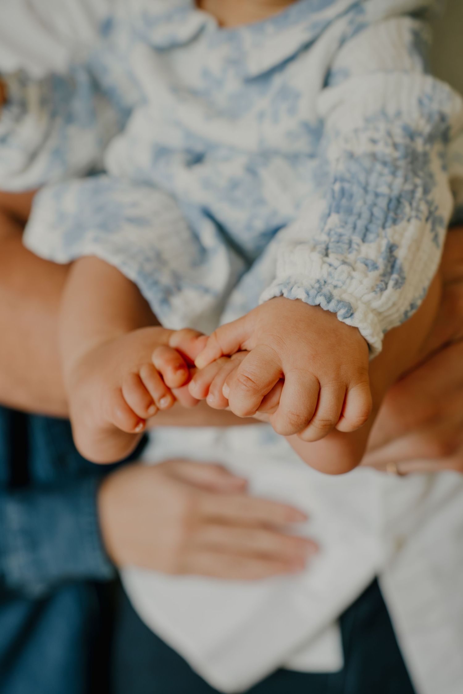 Close-up of tiny newborn baby feet in a blue and white floral outfit, cradled gently in adult hands.