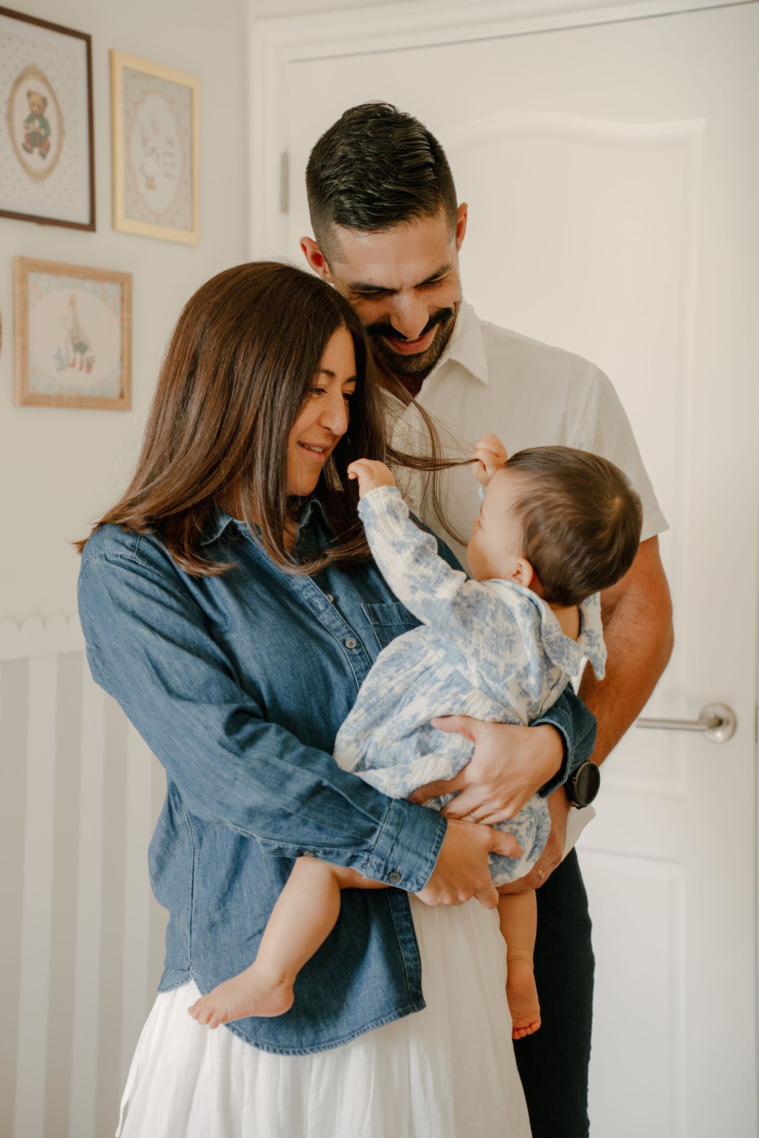A smiling couple in a bright room lovingly holds their baby, dressed in a floral outfit, sharing a joyful family moment.