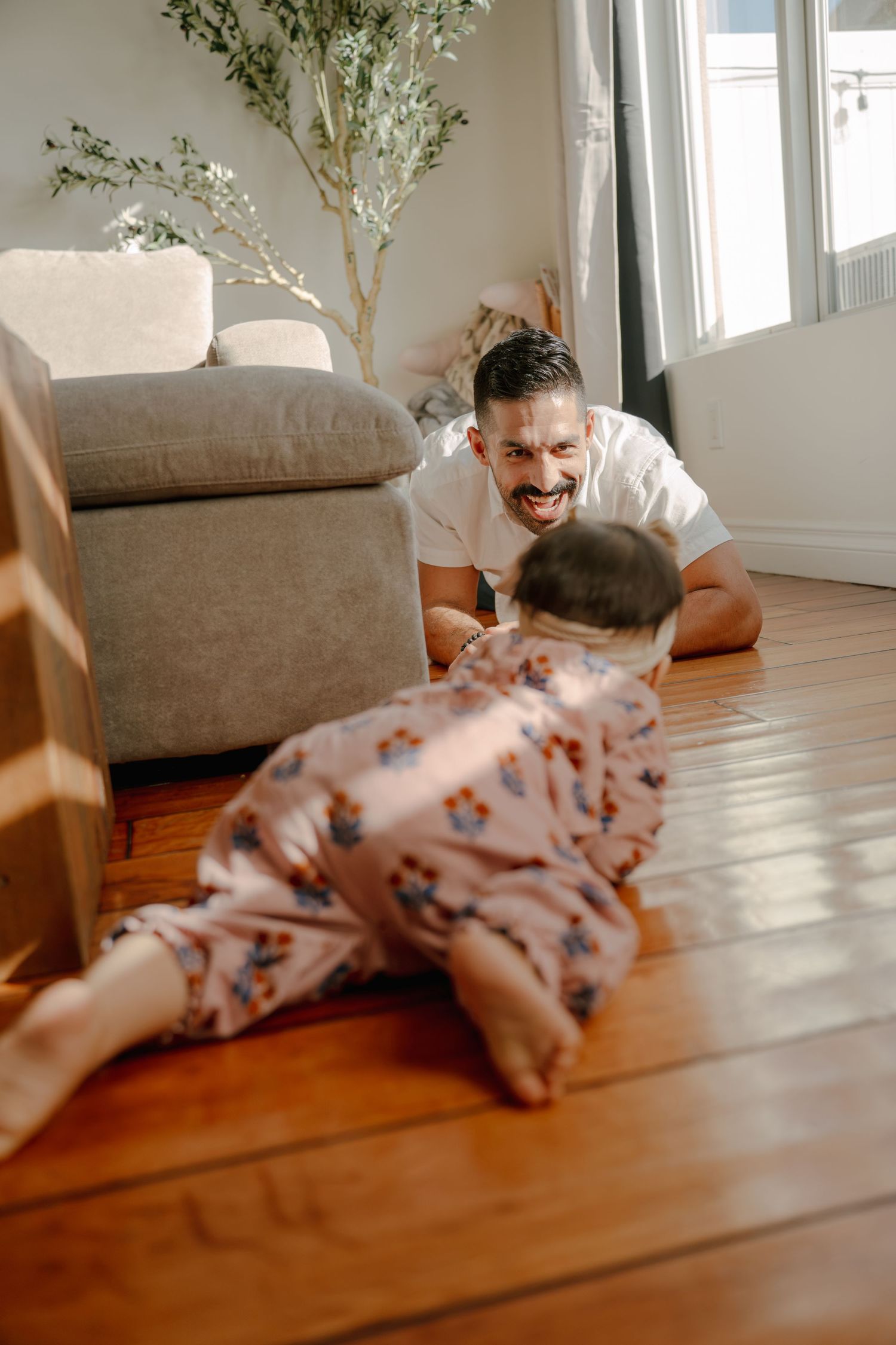 Smiling father lying on hardwood floor playing with crawling baby in floral onesie in bright living room.