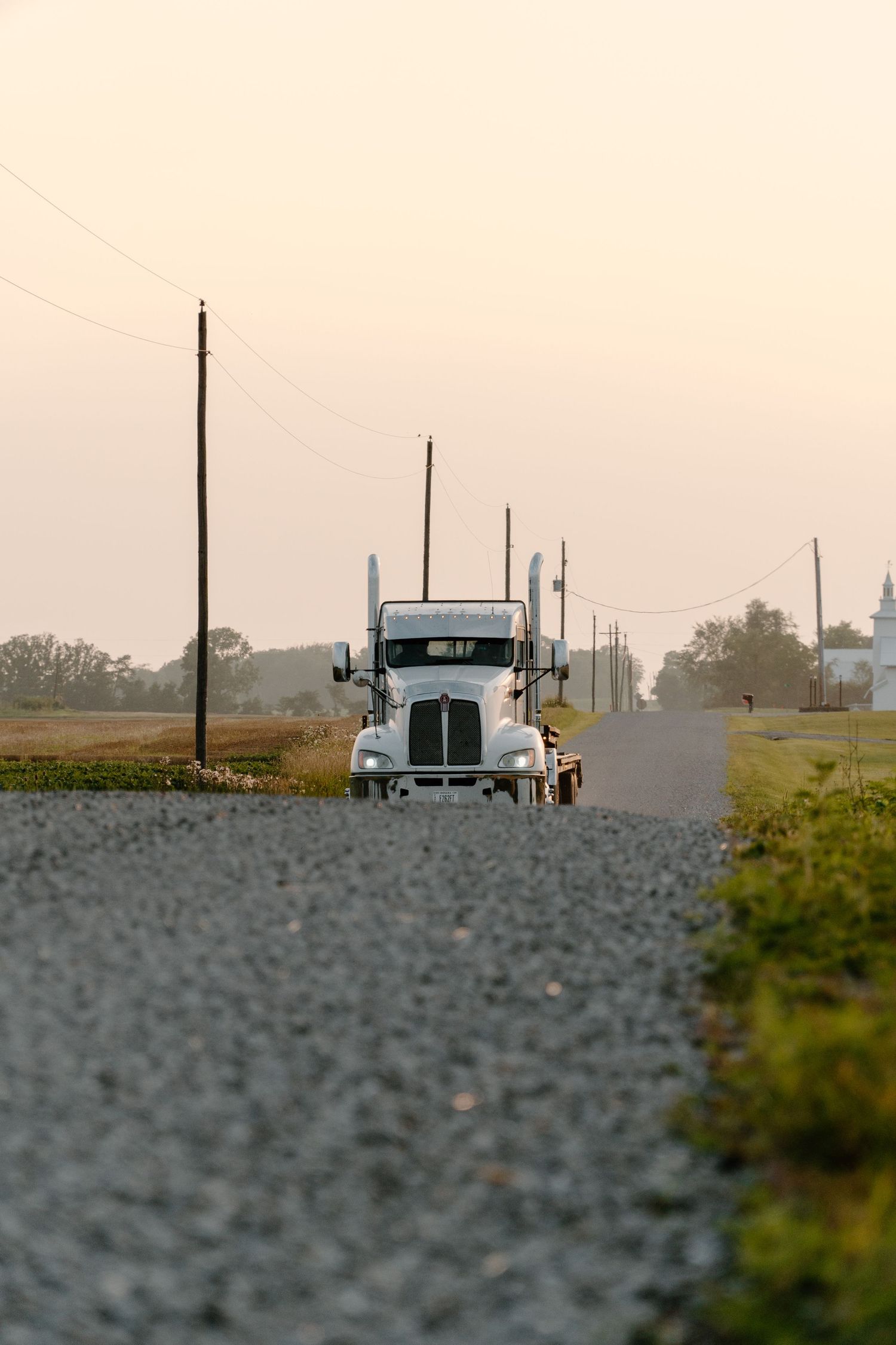 White semi-truck driving toward camera on a rural gravel road at sunset.