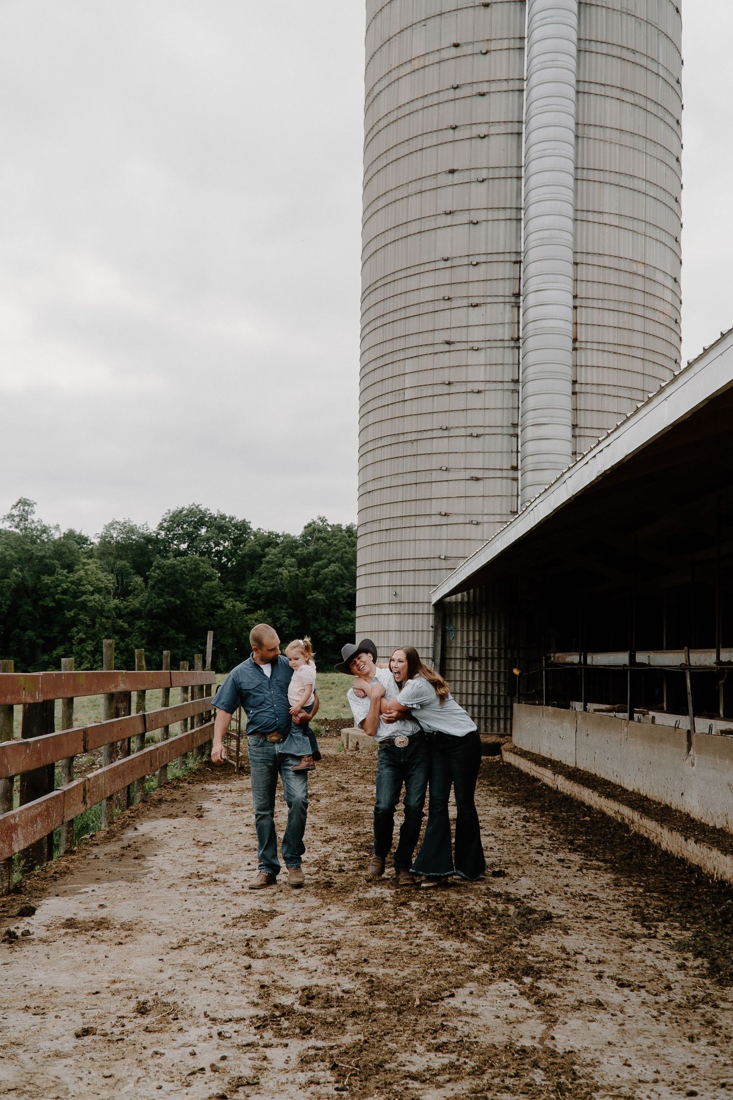 Family of four walking together on muddy farm path beside large grain silo.
