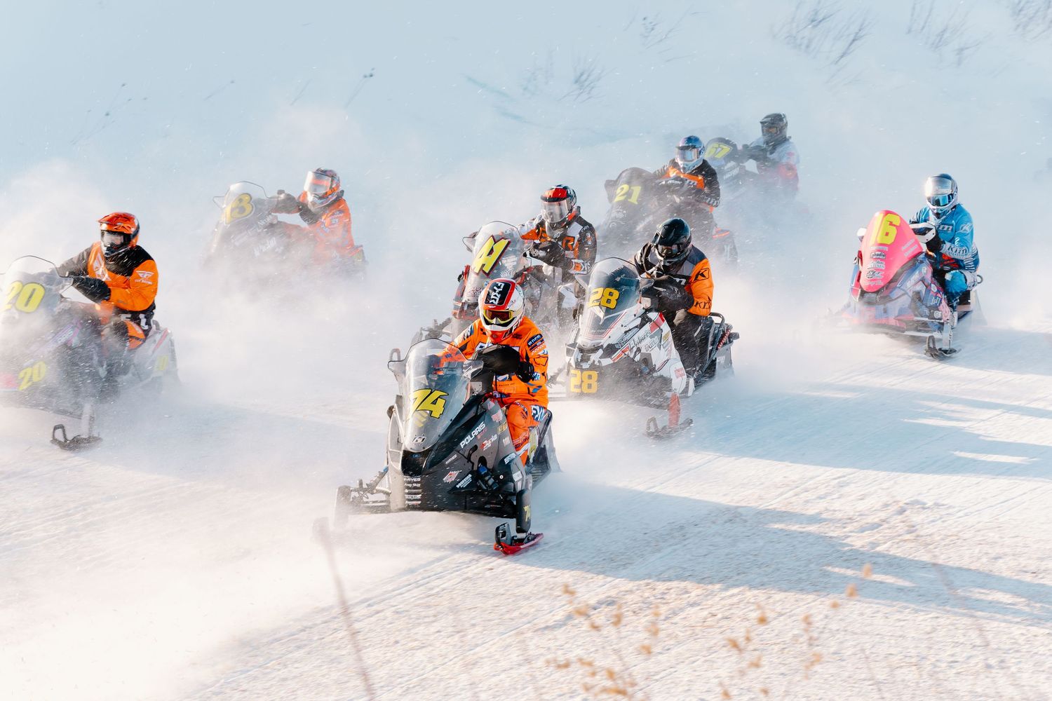 Group of snowmobile racers in colorful gear competing in a winter snocross race kicking up snow.