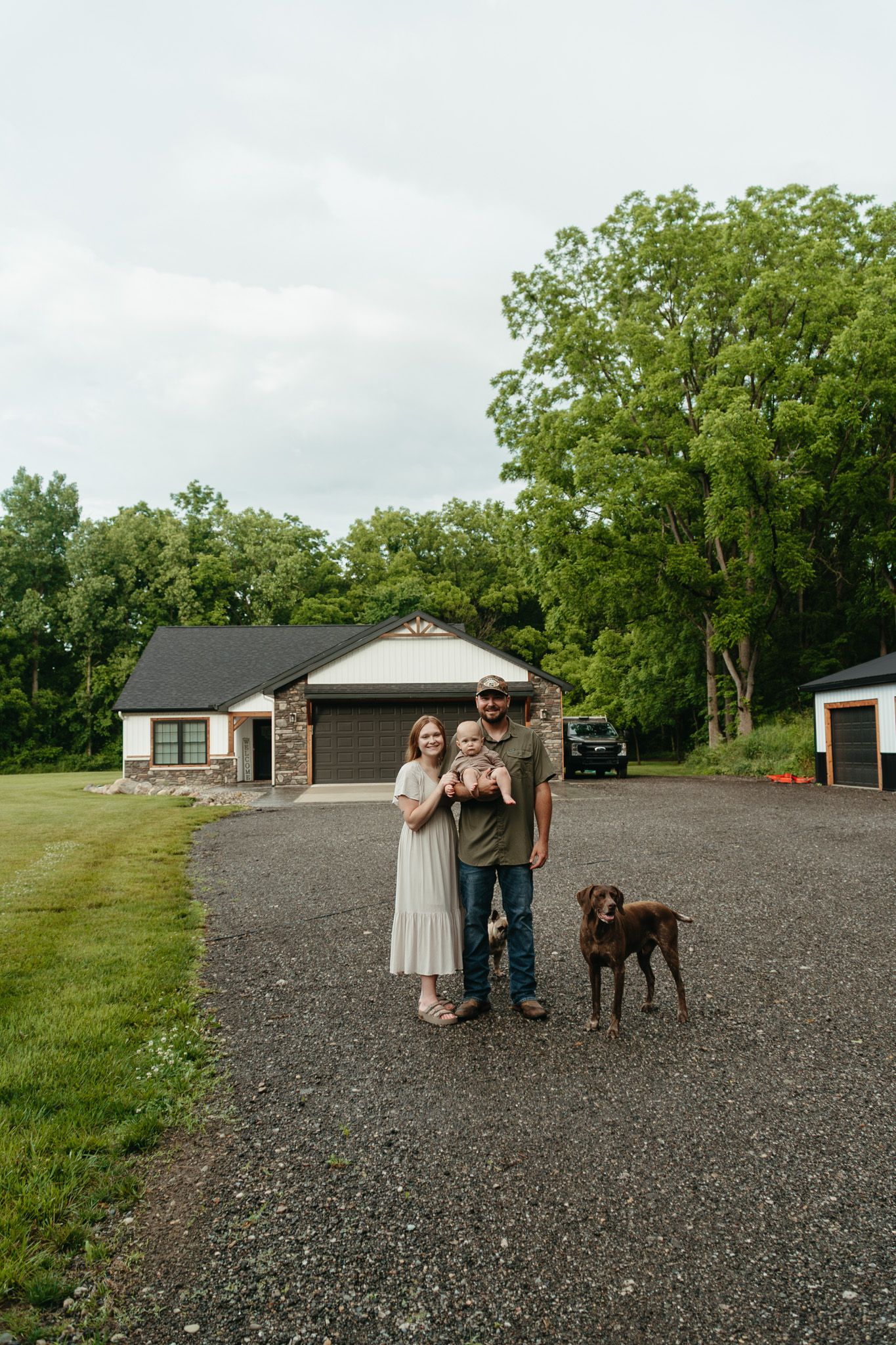 Young family with baby and brown dog posing on gravel driveway in front of ranch home surrounded by green trees.