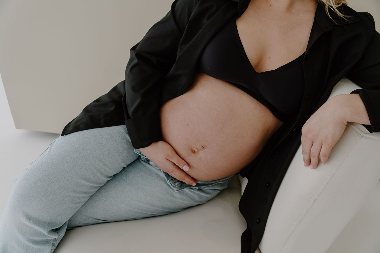 Pregnant woman in black blazer and jeans cradling baby bump while sitting on white couch indoors.