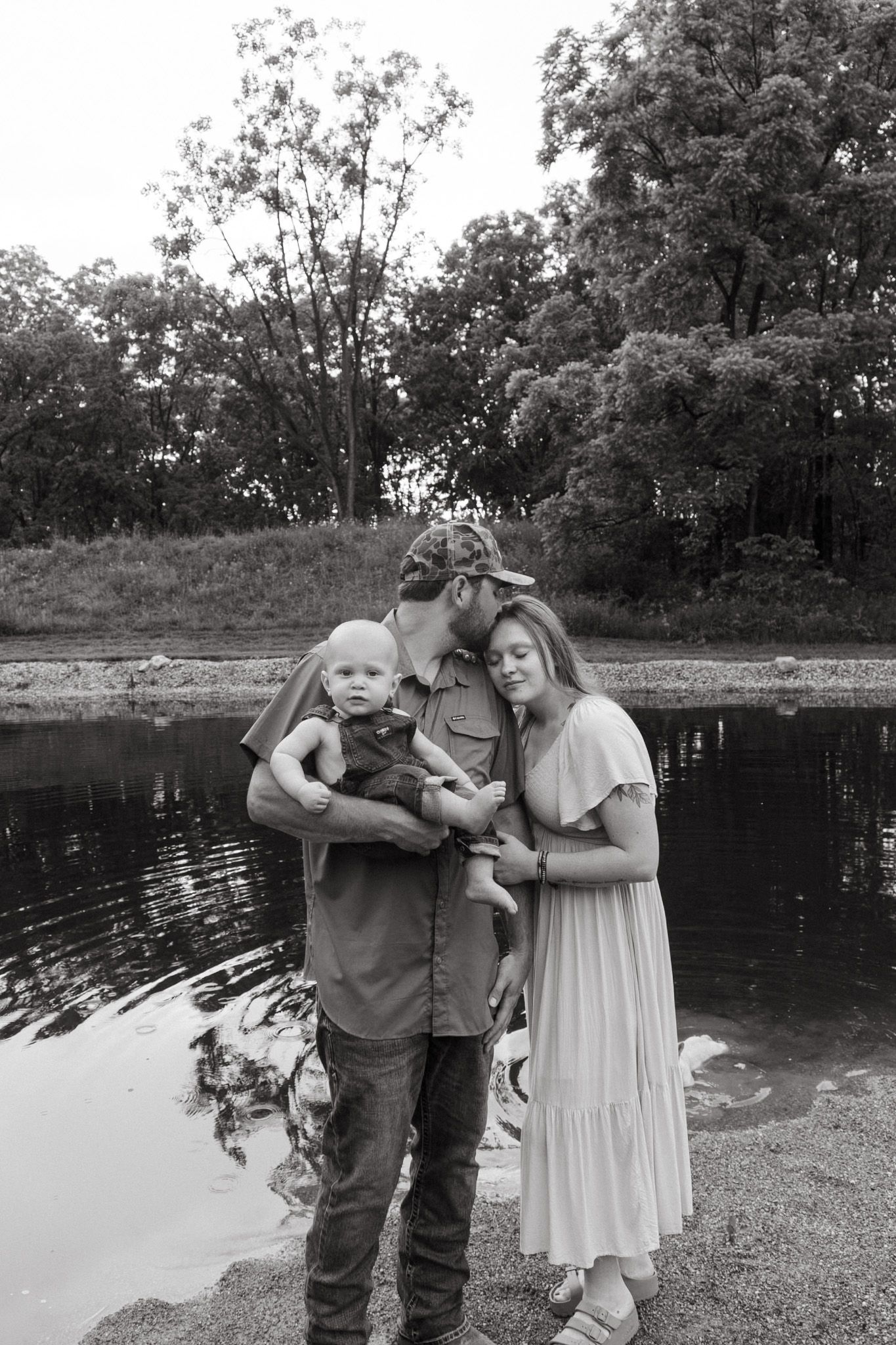 Black and white family portrait of couple with baby standing by peaceful pond surrounded by trees.