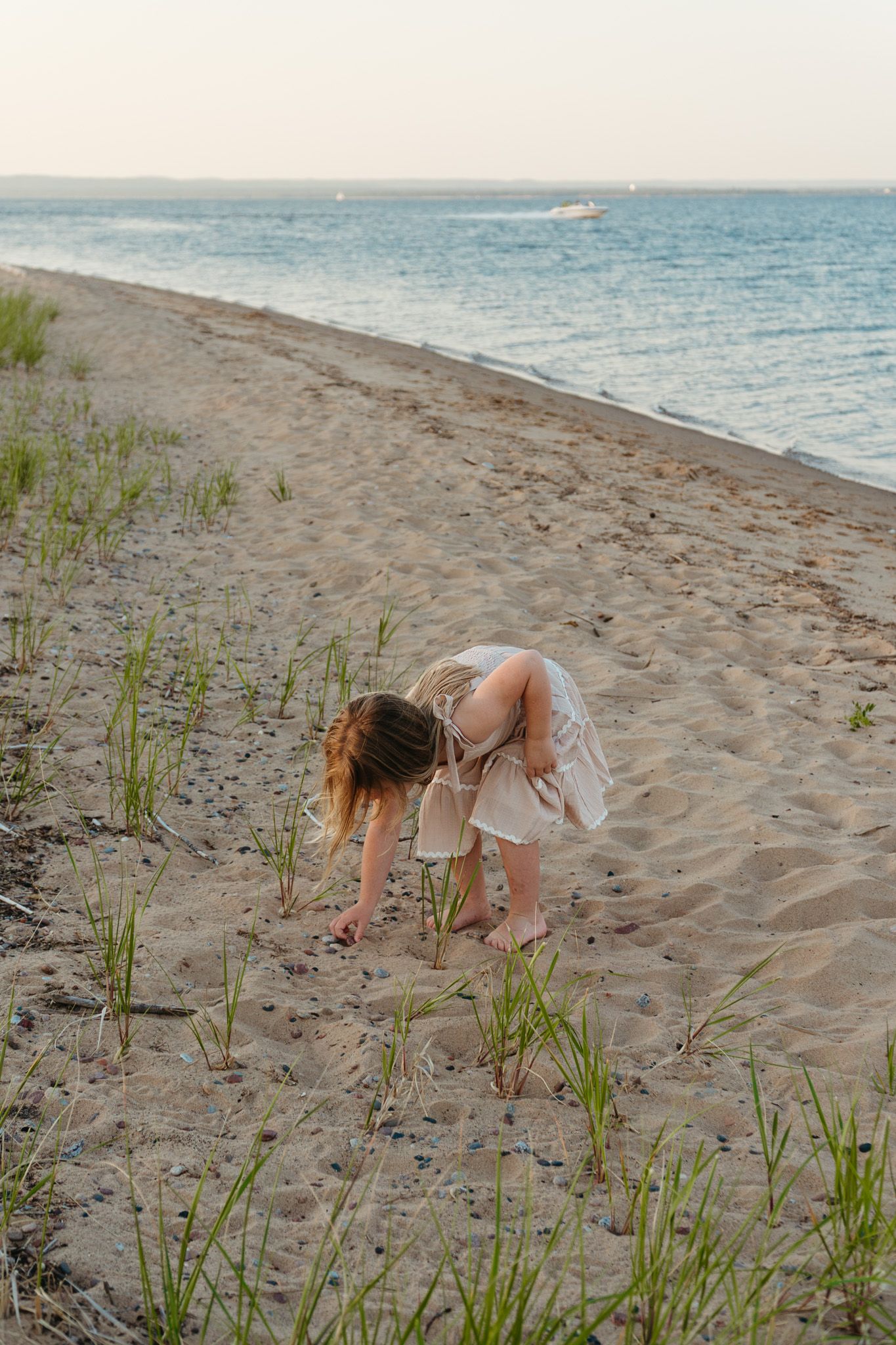 Young girl in white dress bending down exploring beach grass on sandy shoreline at dusk.
