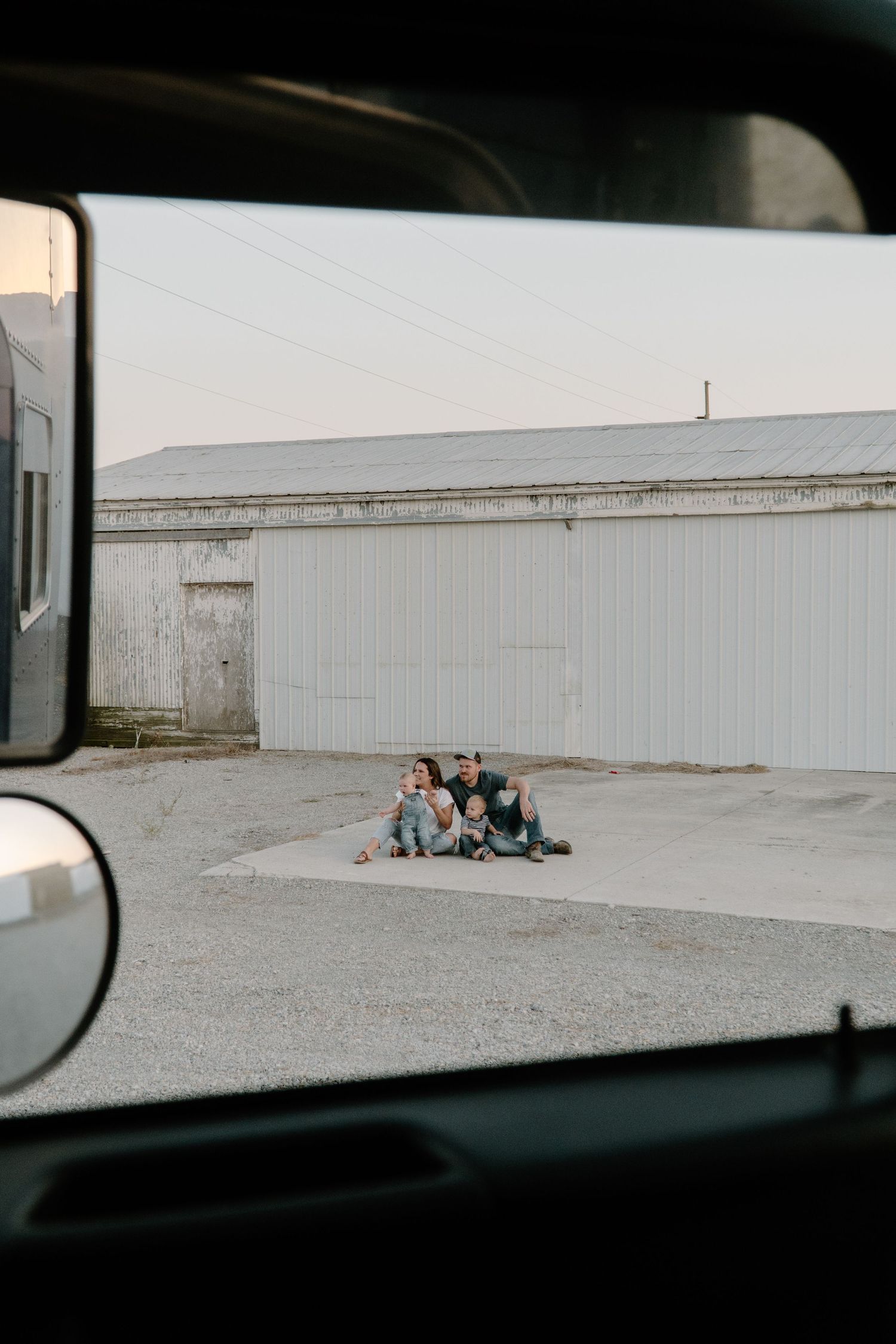 Family of three sitting together on gravel outside a white barn, photographed through a truck window.