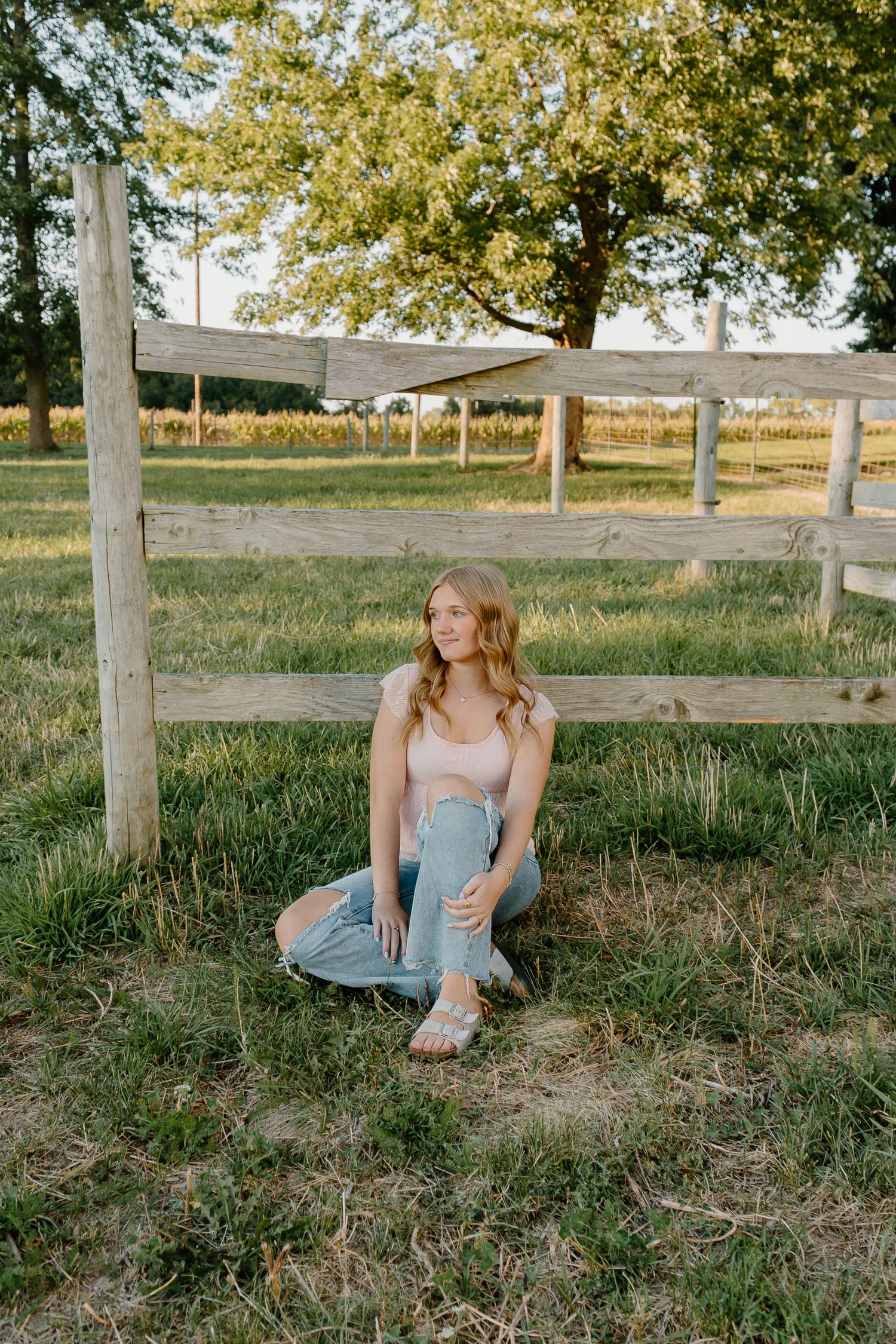 Young woman in a pink top and ripped jeans sitting by a wooden fence in a sunny green pasture.