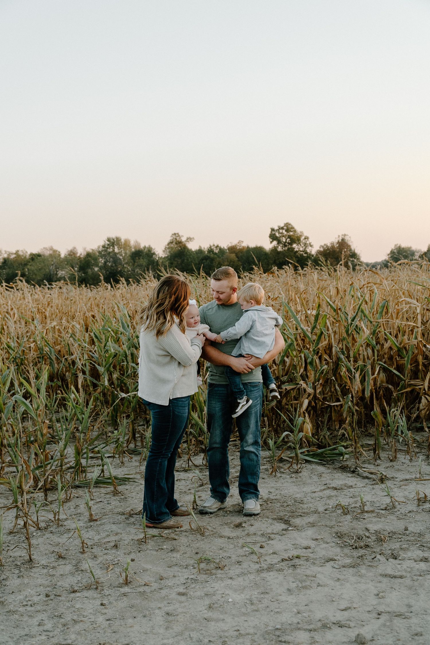 Young family with baby standing in front of a tall green cornfield at dusk.