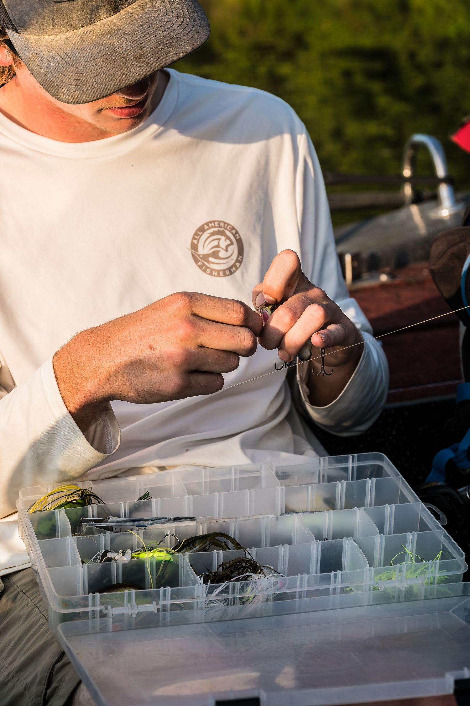 Young man in white shirt selecting fishing lures from organized tackle box at golden hour.