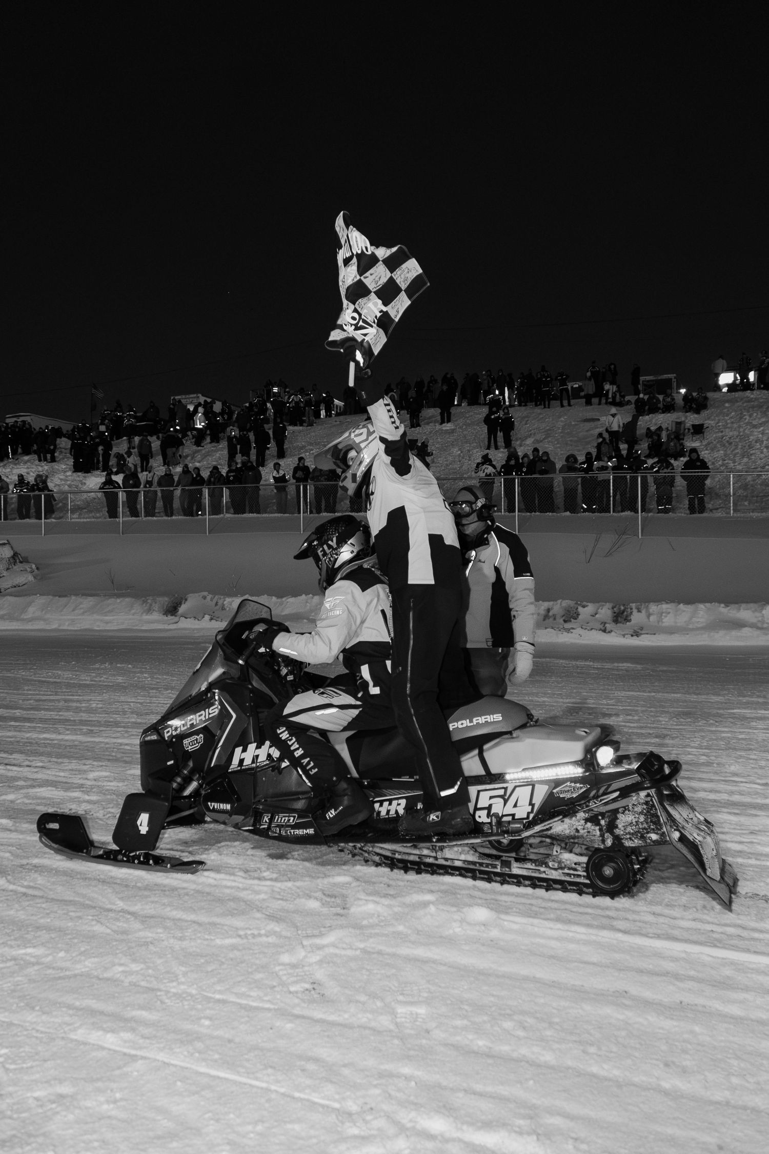 Black and white photo of snowmobile racer number 54 waving checkered flag in victory at a night race.