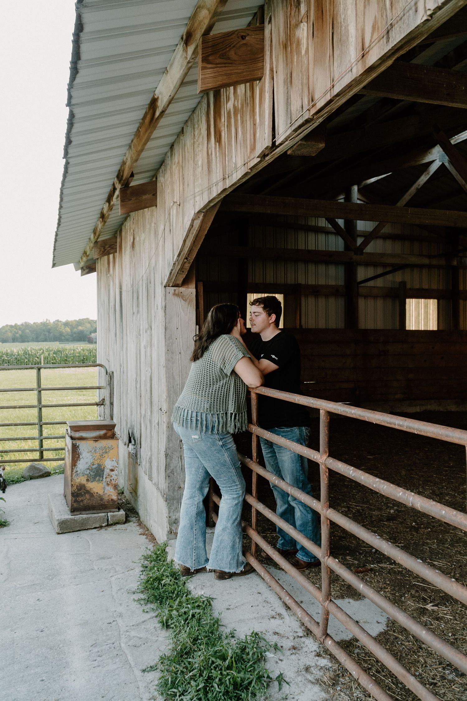 Young couple sharing a kiss at the entrance of a rustic wooden barn with farm fields behind.