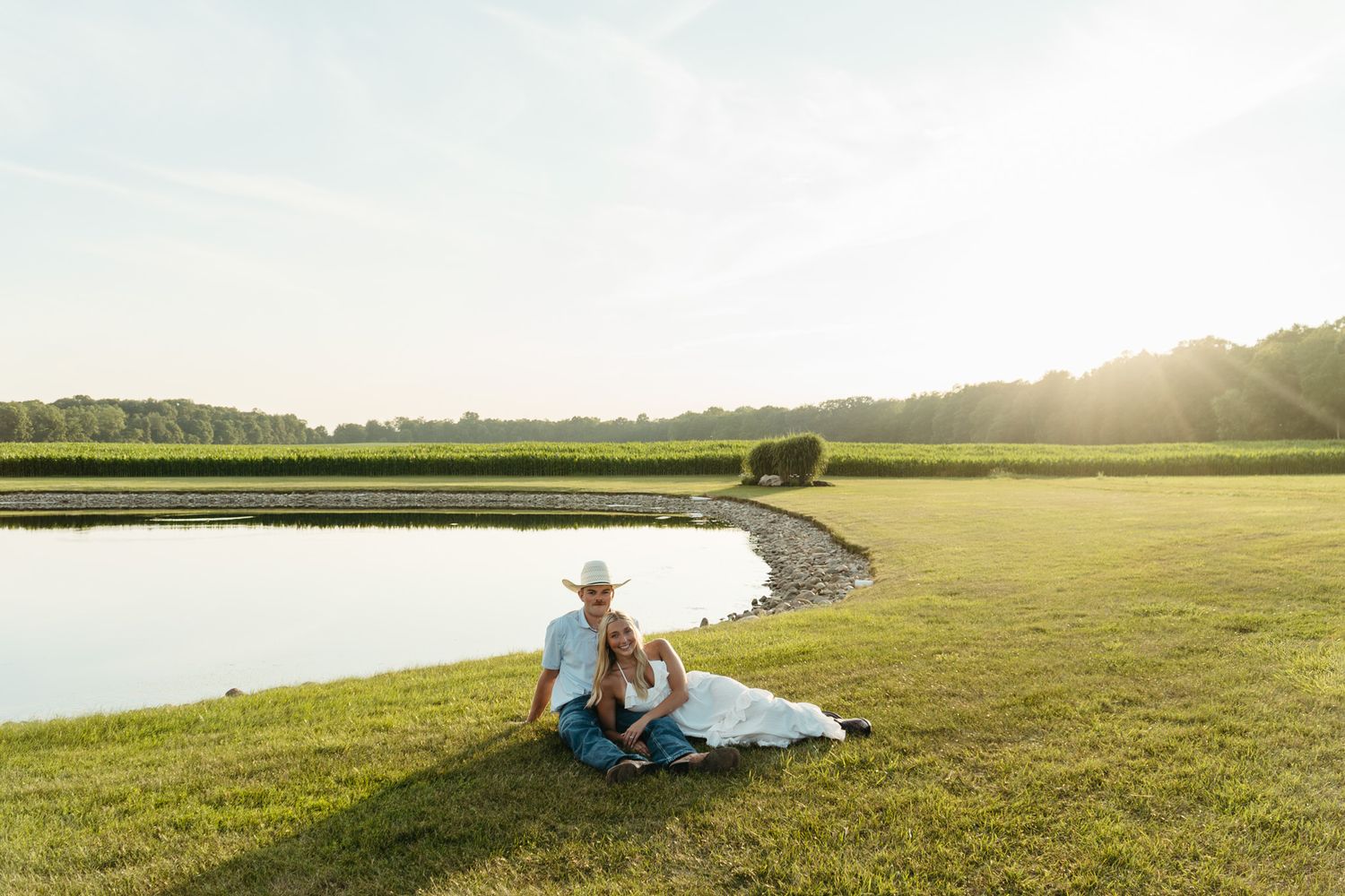 Couple sitting together on grass by a pond at golden hour on a peaceful farm property.