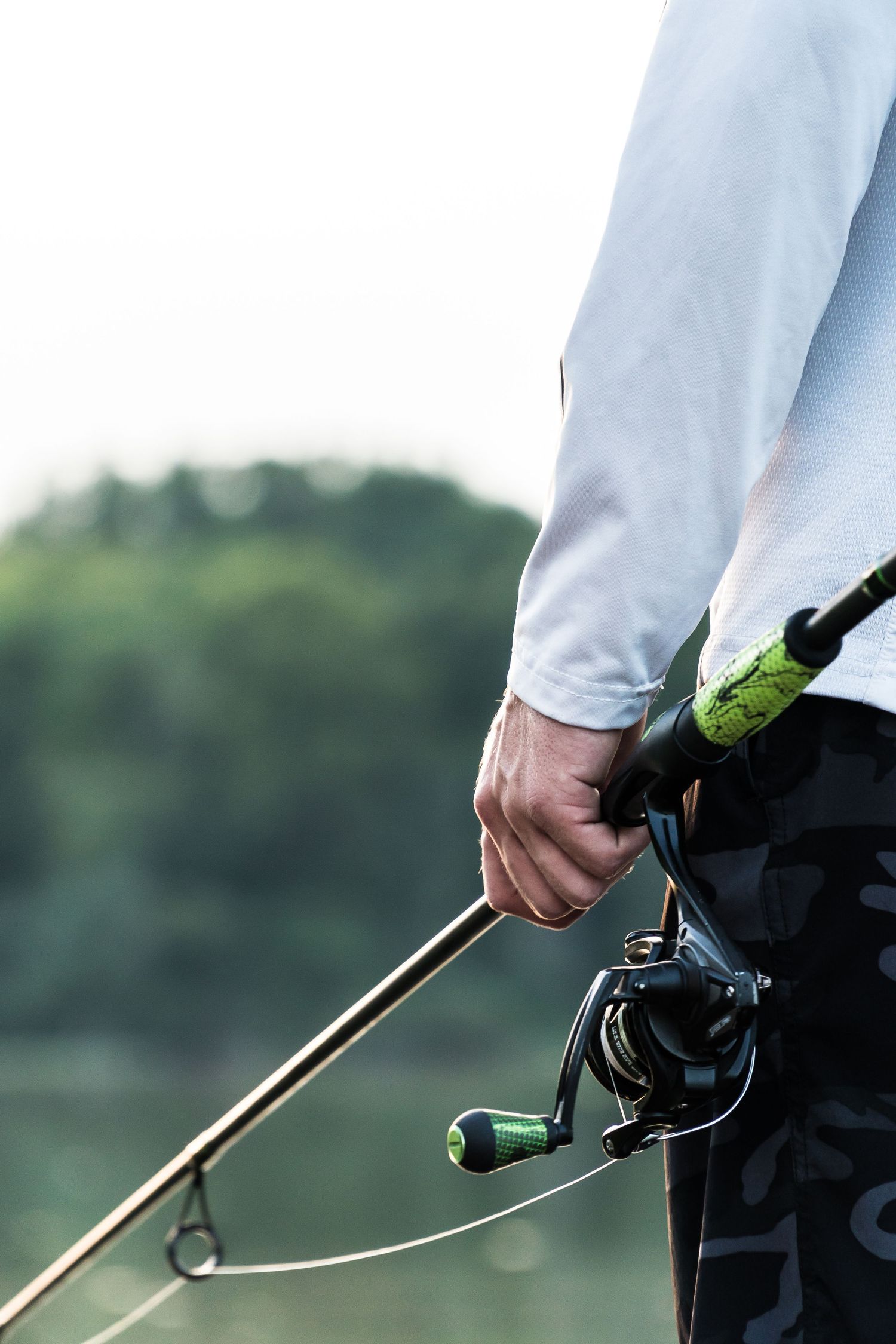 Close-up of fisherman holding baitcasting rod and reel near green tree-lined lake.