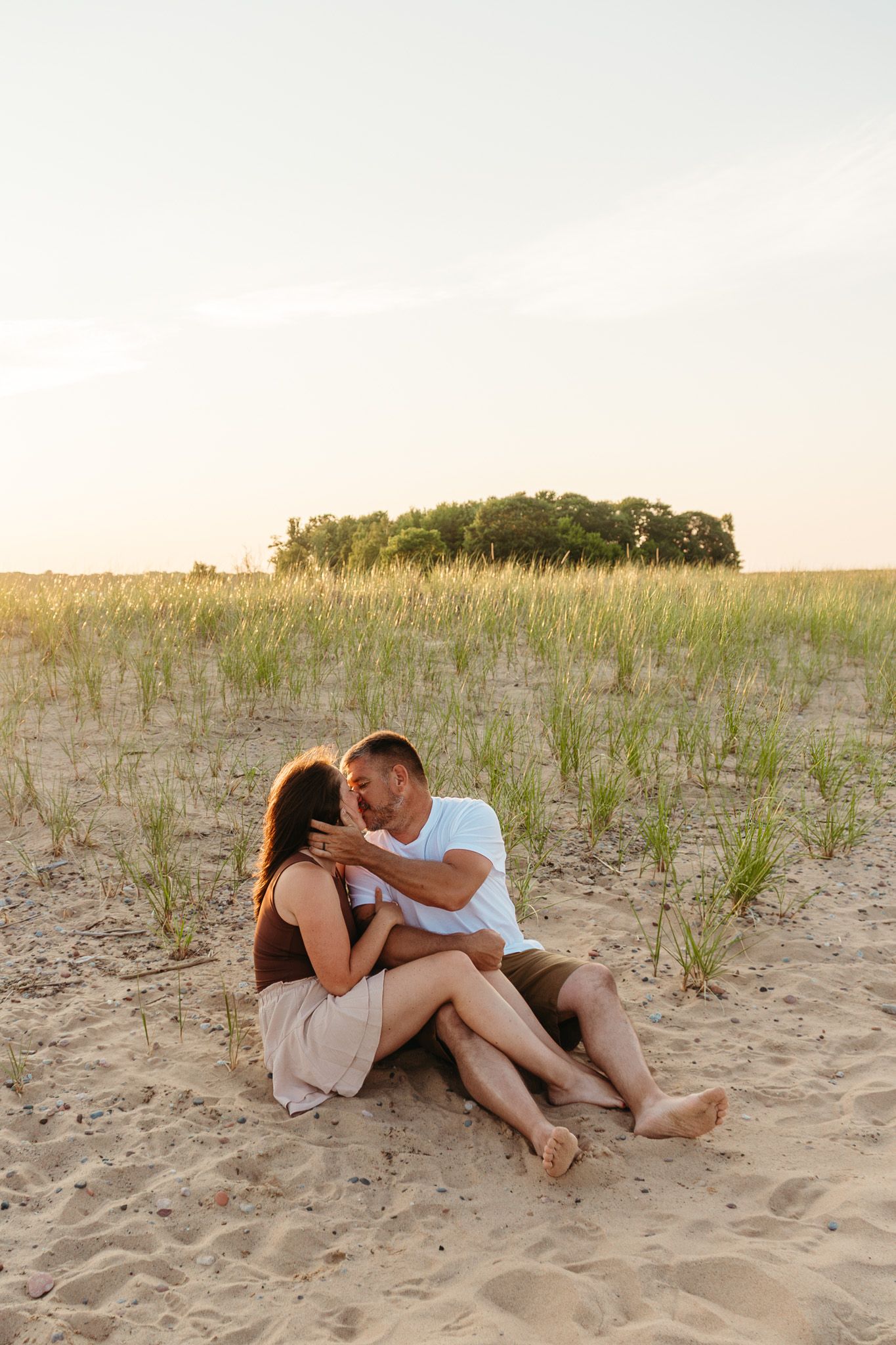 Romantic couple sitting together on sandy beach dunes at golden hour, sharing an intimate moment.