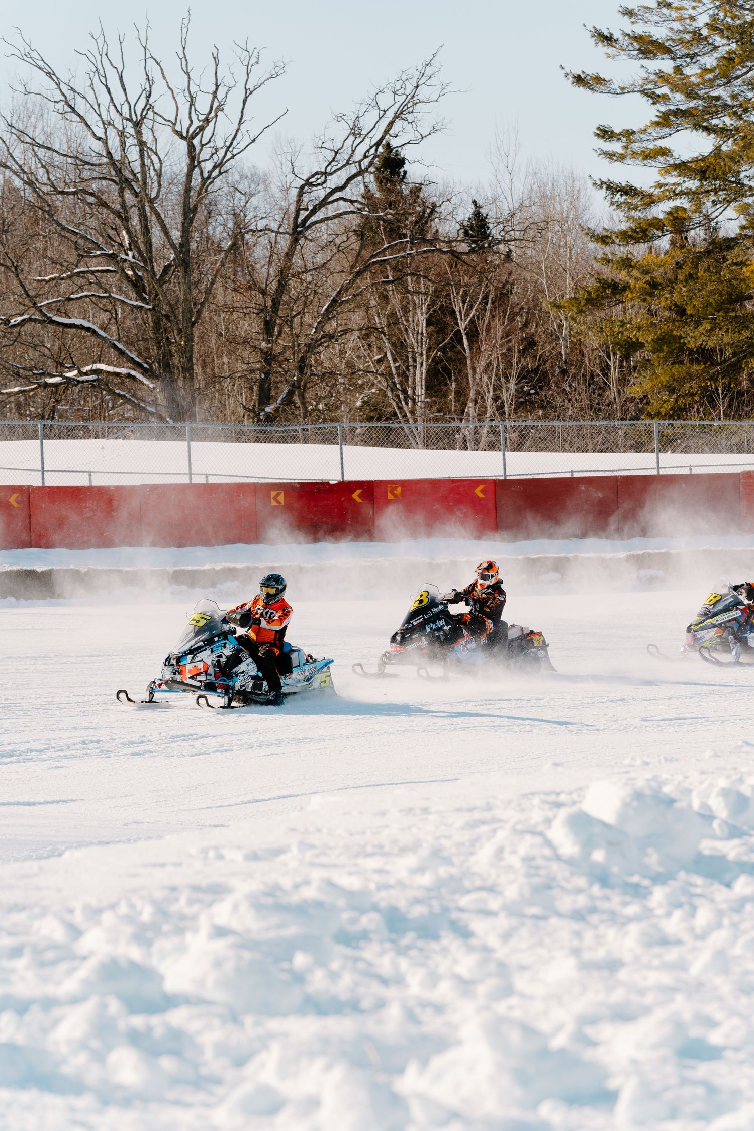 Three snowmobile racers competing on a snowy track with red barriers and snow-covered trees in background.