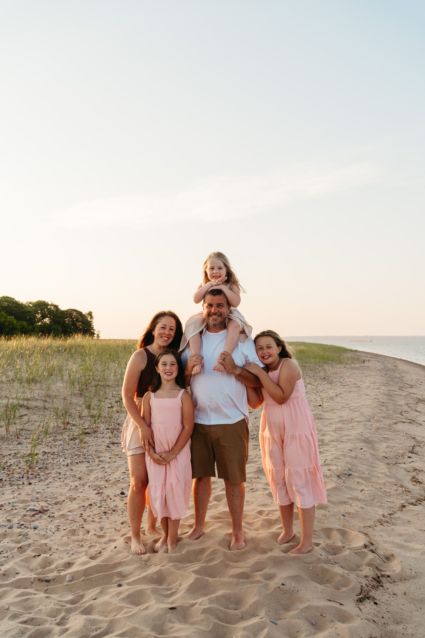 Happy family of five posing on sandy beach at sunset, father carrying youngest child on shoulders.