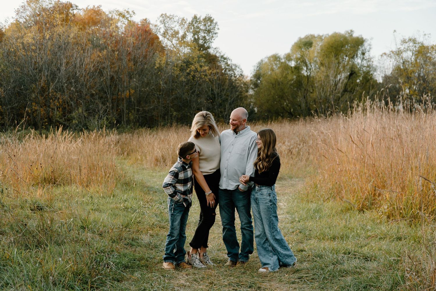 Family of four standing together on a path in an autumn field with tall golden grass.