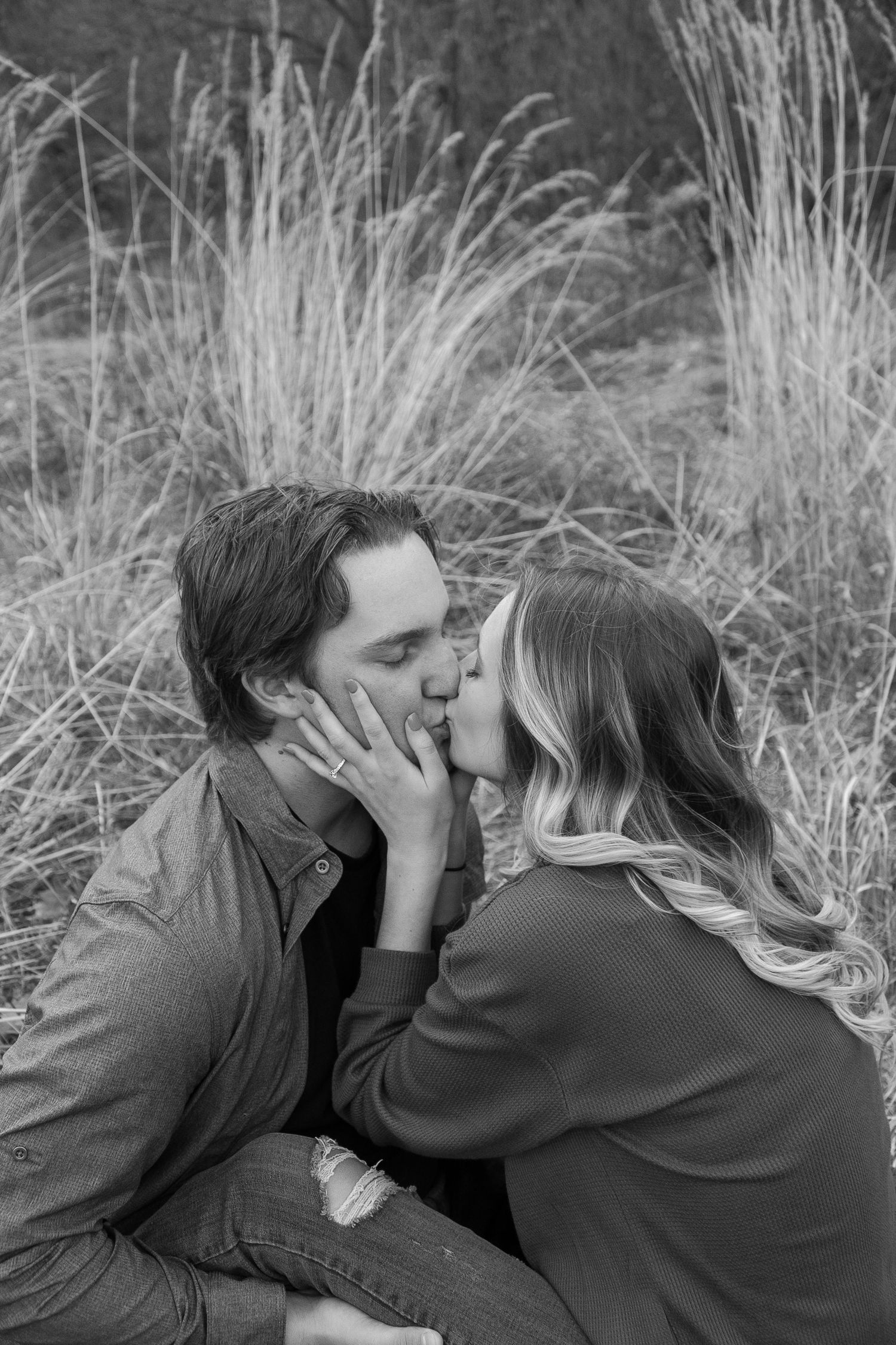Black and white photo of a couple sharing a romantic kiss outdoors among tall grass.