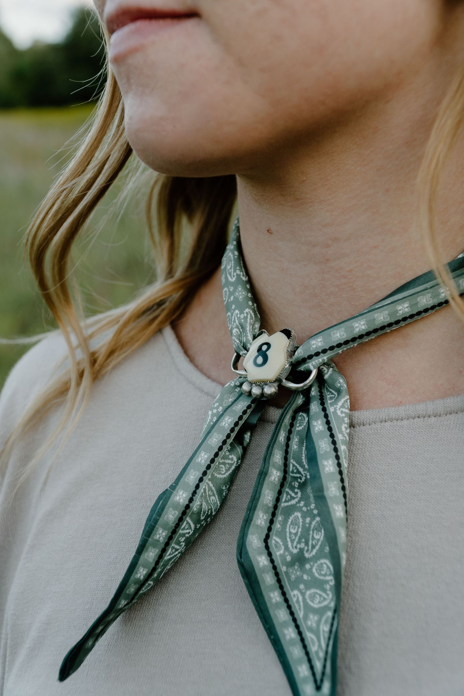 Close-up of woman wearing green paisley bandana scarf tied with a vintage number 8 pin.