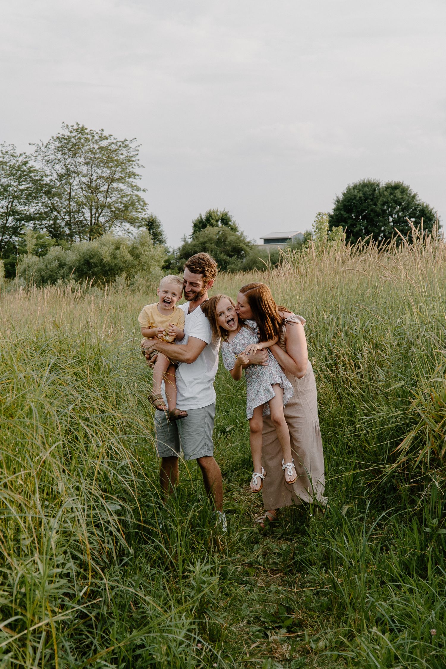 Happy family of four laughing together in tall grass meadow on overcast day.