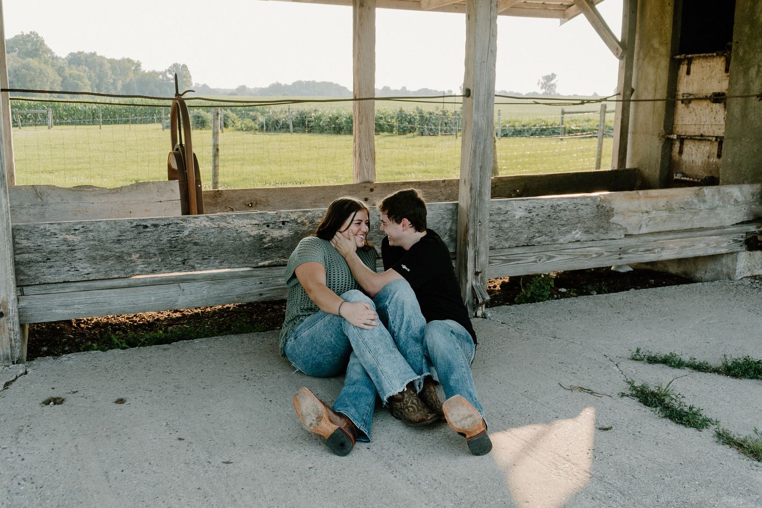 Couple sitting together leaning against rustic barn wall with green farm fields in background.