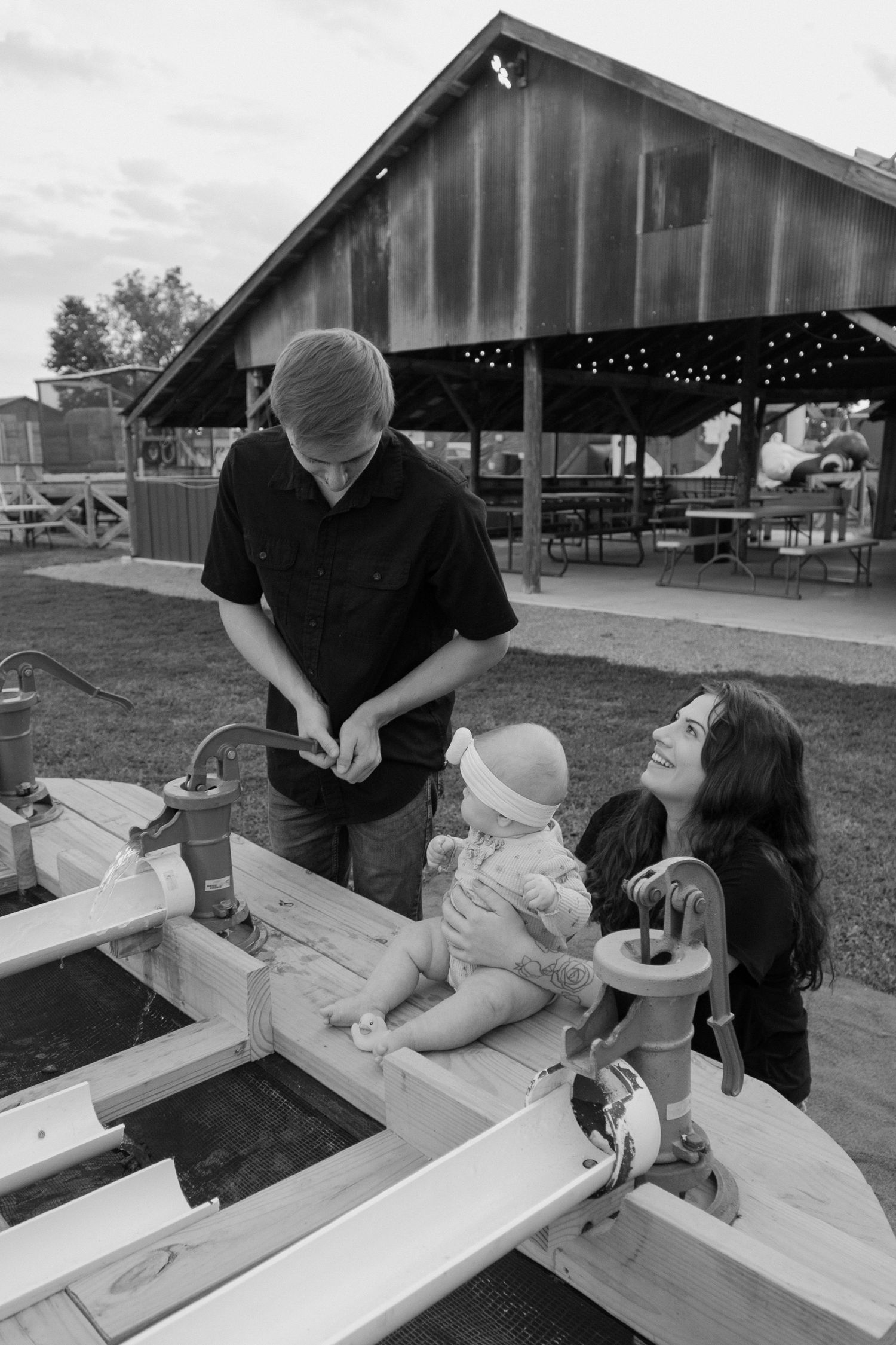 Black and white photo of a young family with a baby near a wooden structure at a rustic venue.