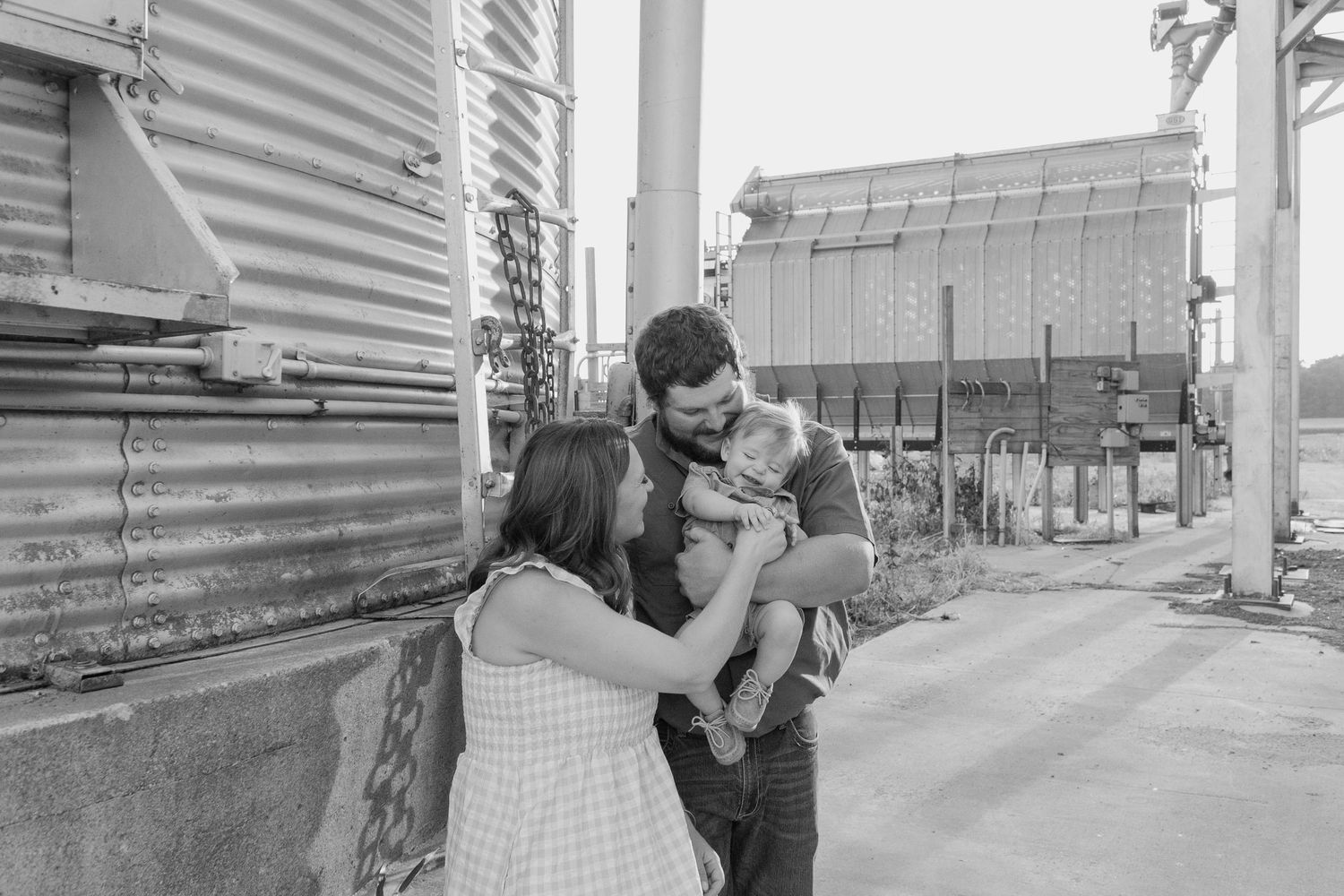 Black and white photo of couple sharing tender moment with baby at industrial farm setting.