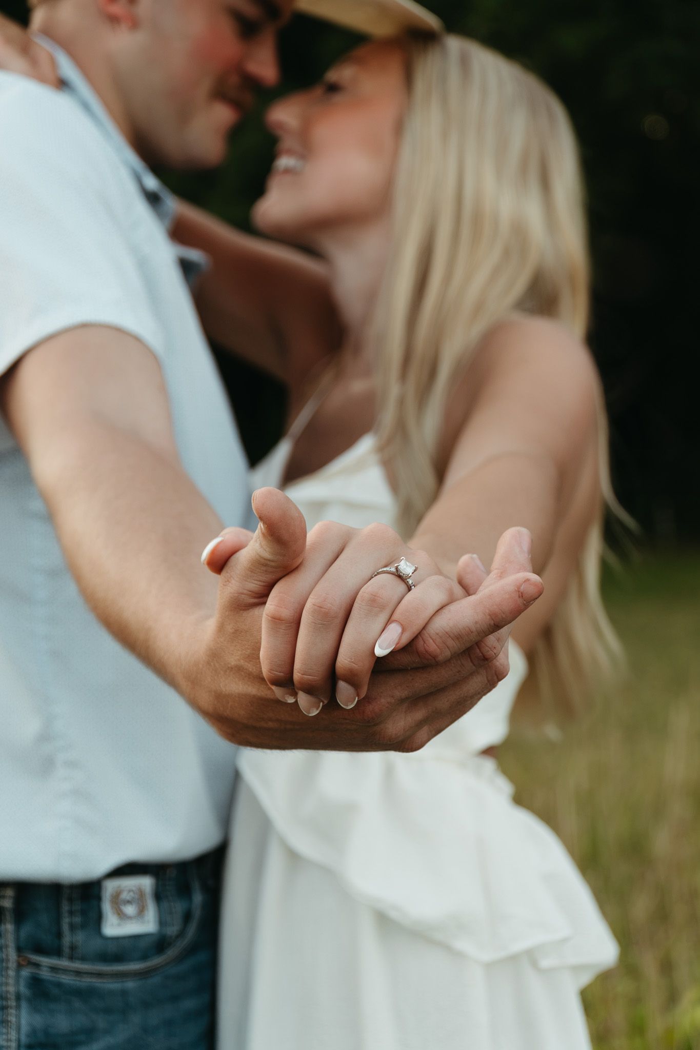 Couple holding hands showing engagement ring while almost kissing outdoors.