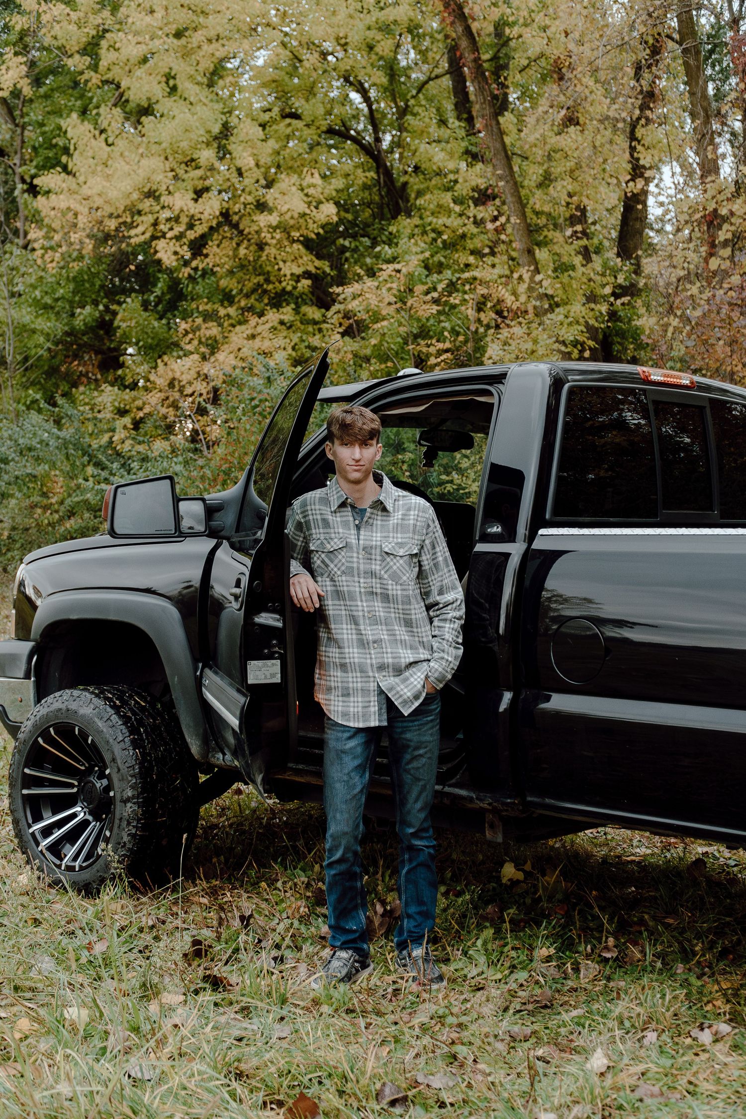 Teen boy in plaid shirt standing beside open door of lifted black GMC truck in fall foliage setting.