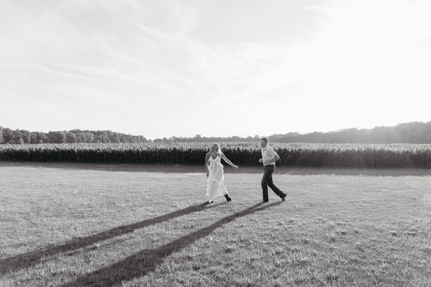 Black and white photo of couple holding hands while walking through an open field near a cornfield.